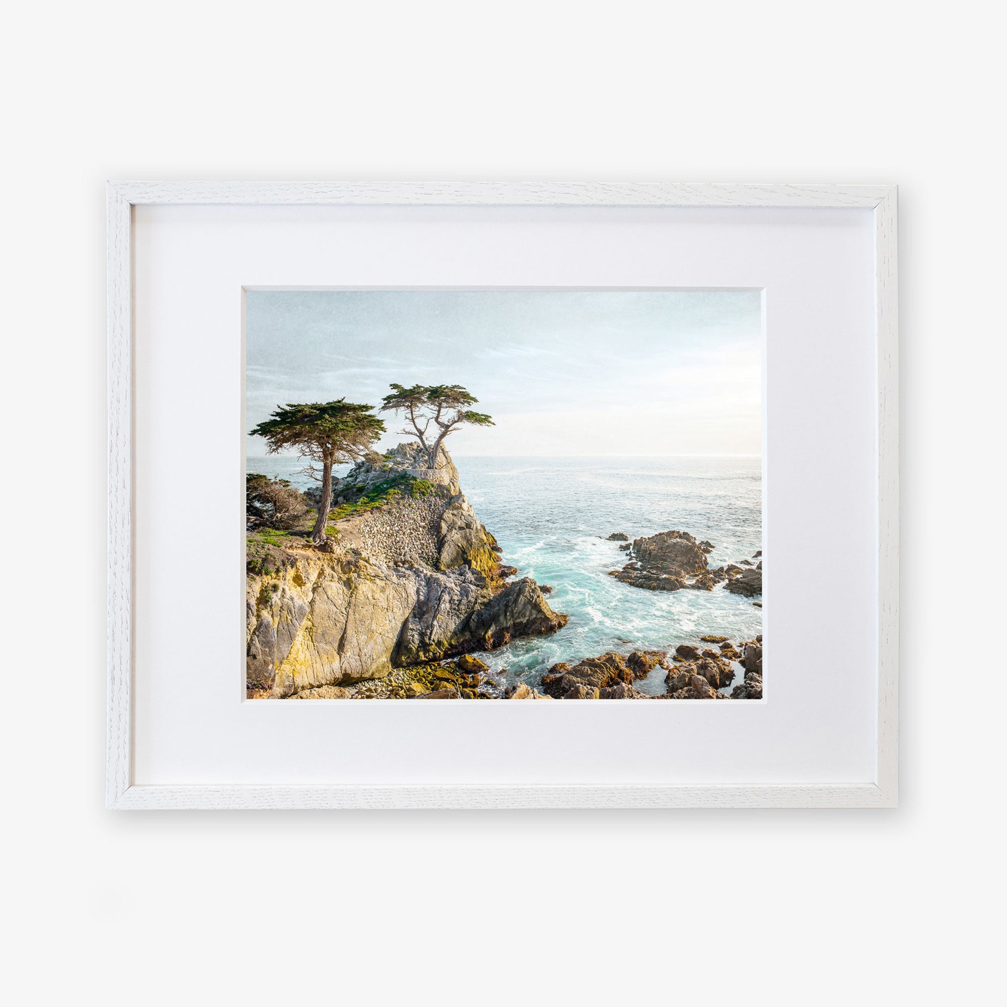 A framed painting of a California Coastal Print, &#39;Lone Cypress&#39; with rugged cliffs, choppy sea, and windswept trees under a bright sky at Pebble Beach, displayed on a white background by Offley Green.