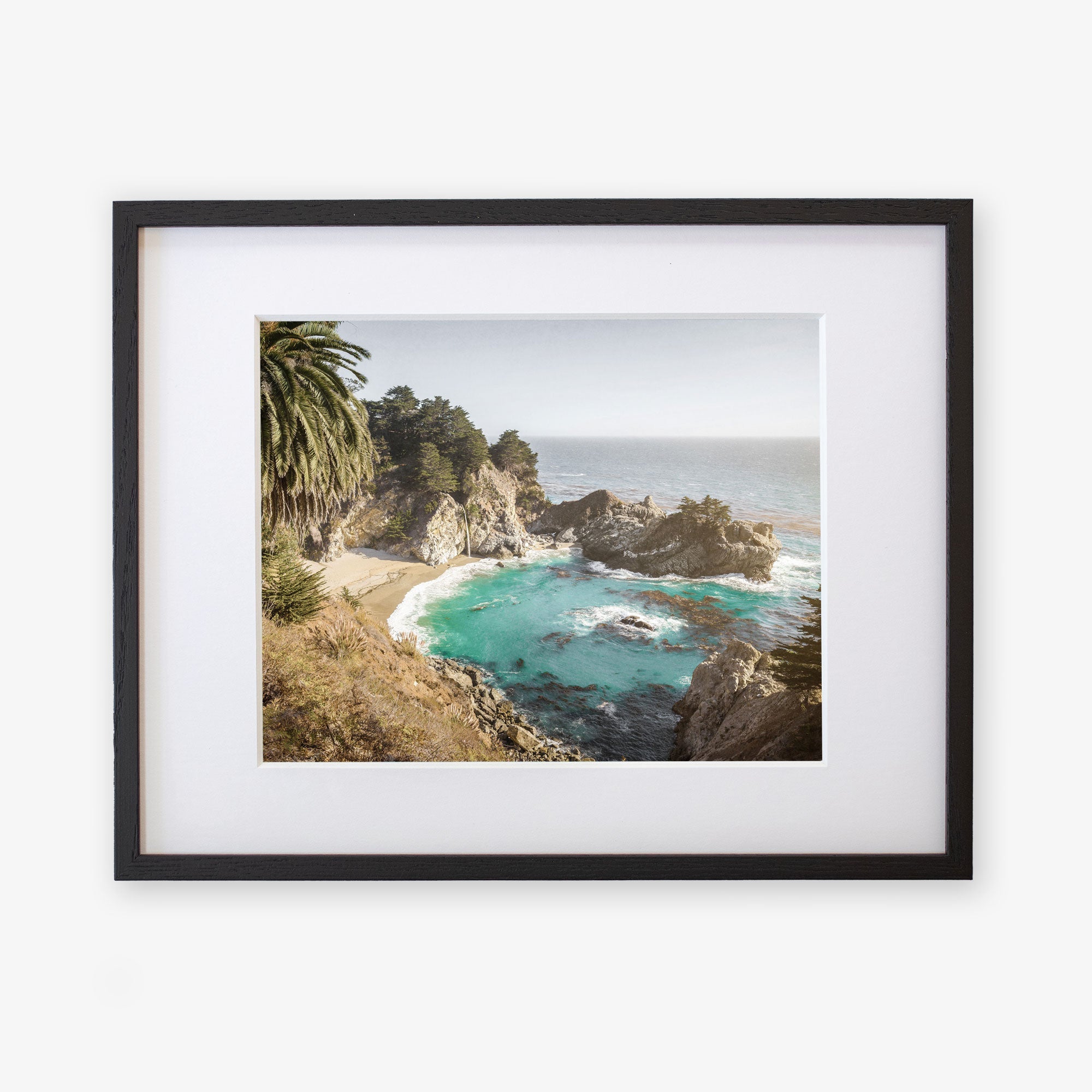 Framed photograph of a Big Sur Coastal Print &#39;Julia Pffeifer&#39; wall art view with rocky cliffs and turquoise waters, surrounded by green foliage and a sandy beach by Offley Green.