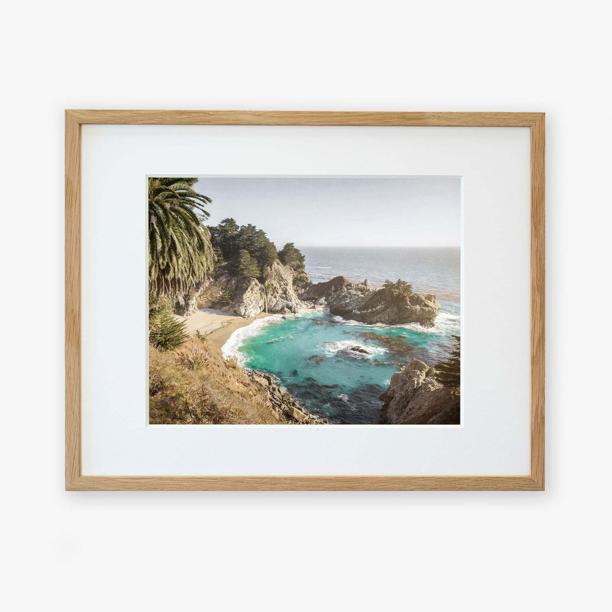 A framed photograph of a Big Sur Coastal Print, 'Julia Pffeifer', featuring a rocky shoreline, azure waters, and lush green foliage under a blue sky by Offley Green.