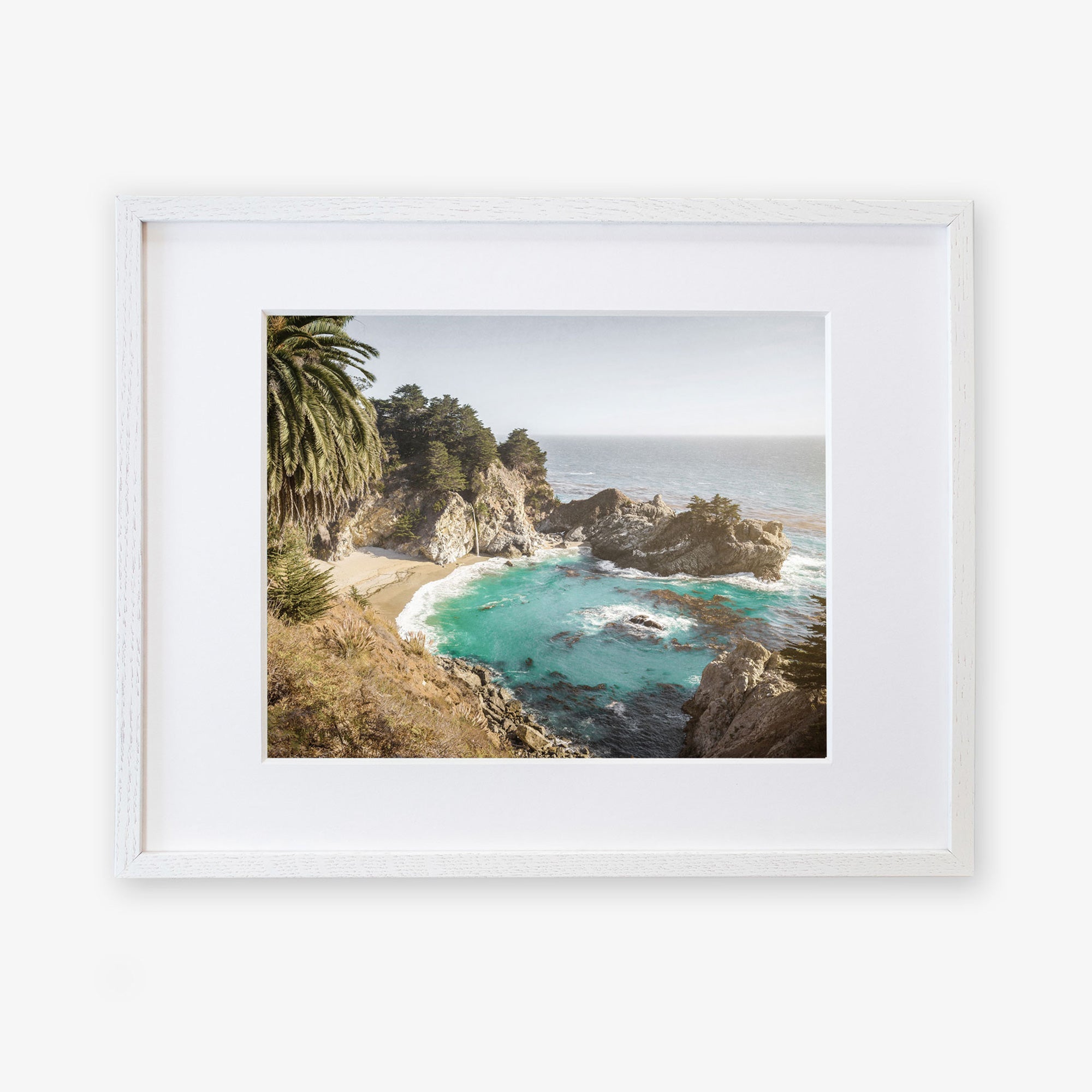 A framed artwork depicting a scenic view of the Pacific Coast Highway with turquoise waters, rocky cliffs, and lush greenery, displayed on a white background is the Big Sur Coastal Print by &#39;Julia Pffeifer&#39; from Offley Green.