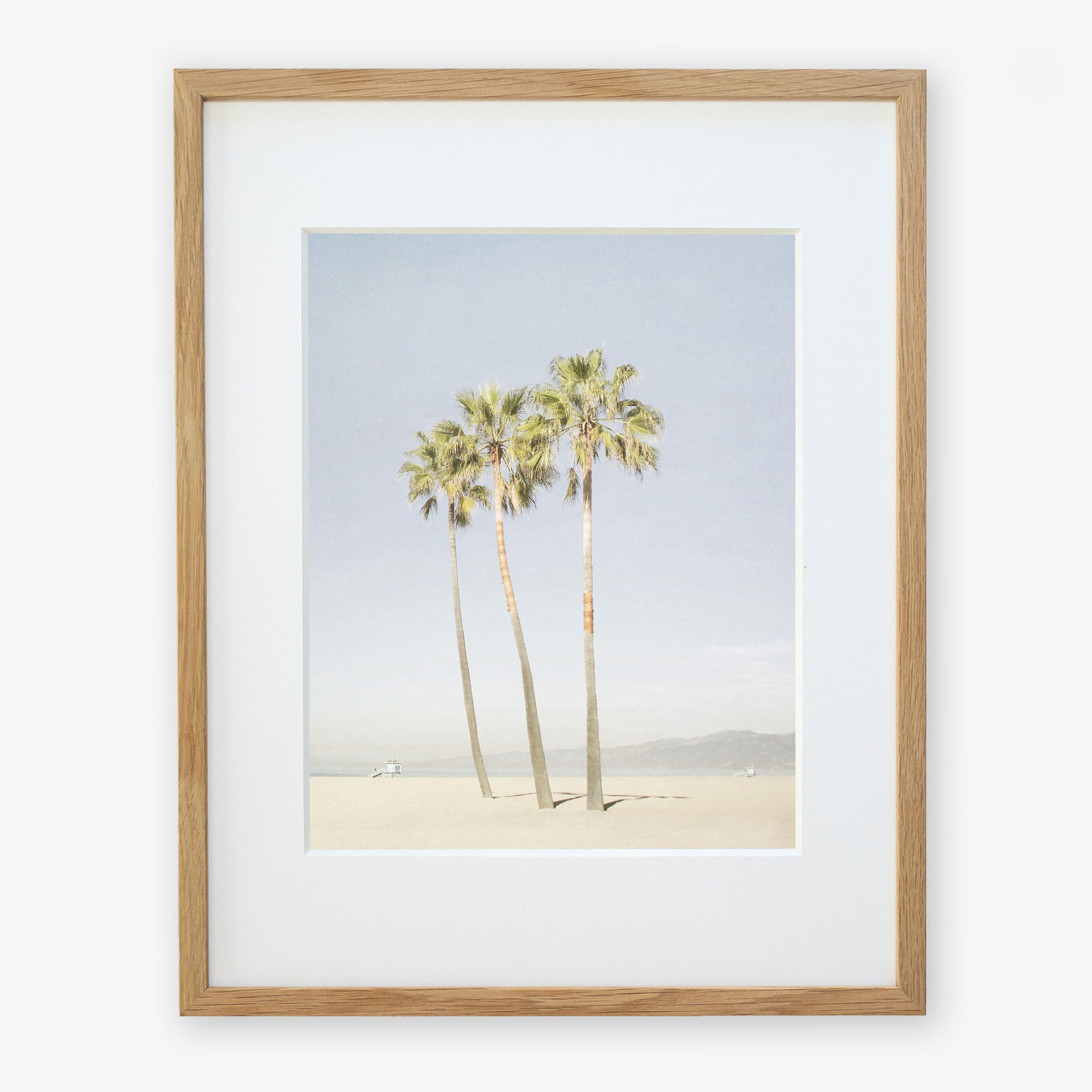 Framed artwork depicting three tall palm trees under a clear sky, with a small figure and a dog at the base, possibly on Venice Beach. The frame is simple and made of light wood. This is the Offley Green California Venice Beach Print, 'Three Palms'.
