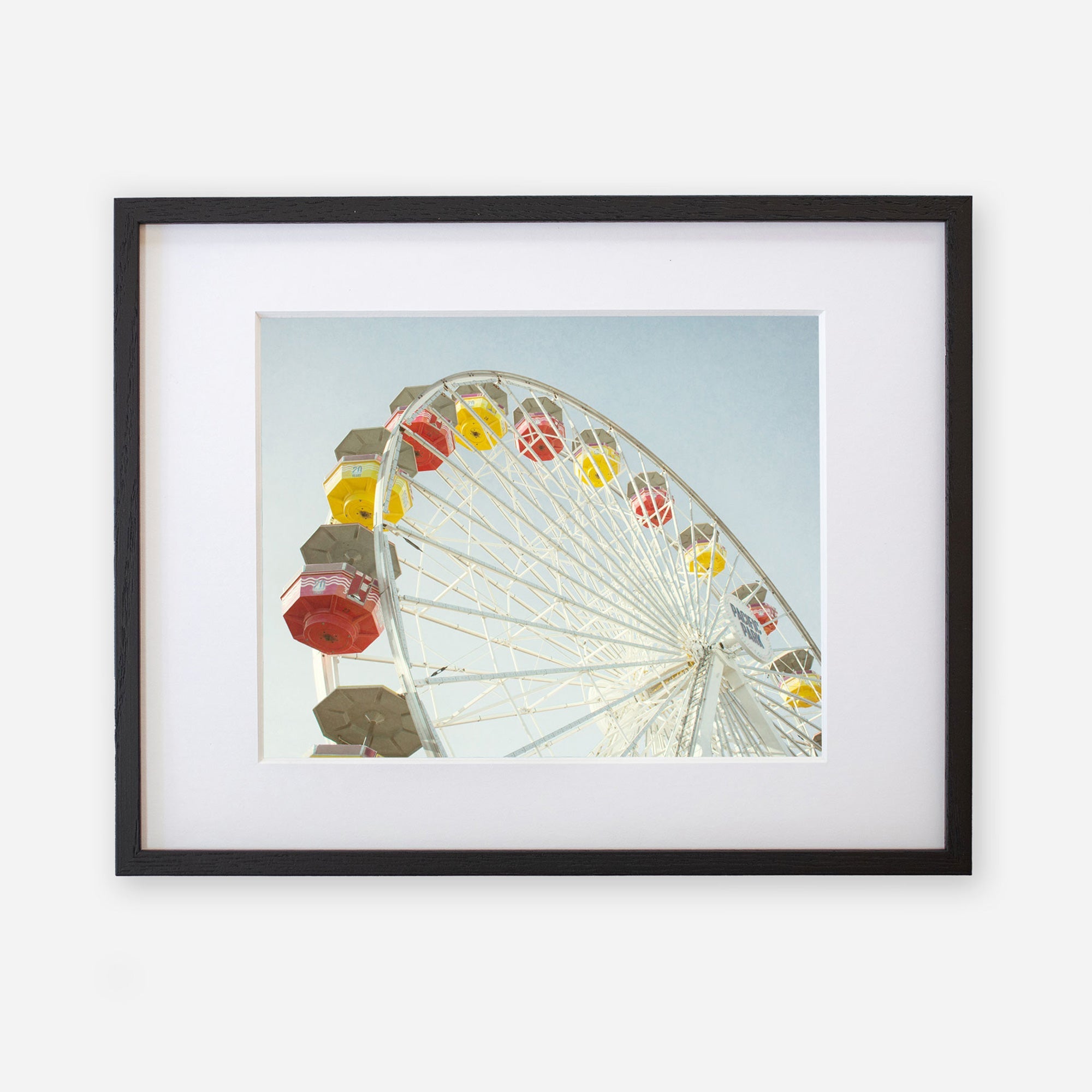 A framed photograph of a colorful Santa Monica Ferris Wheel Print, &#39;Ferris Above&#39;, printed on archival photographic paper, featuring brightly colored cabins in yellow, red, and orange against a clear sky. Brand: Offley Green.