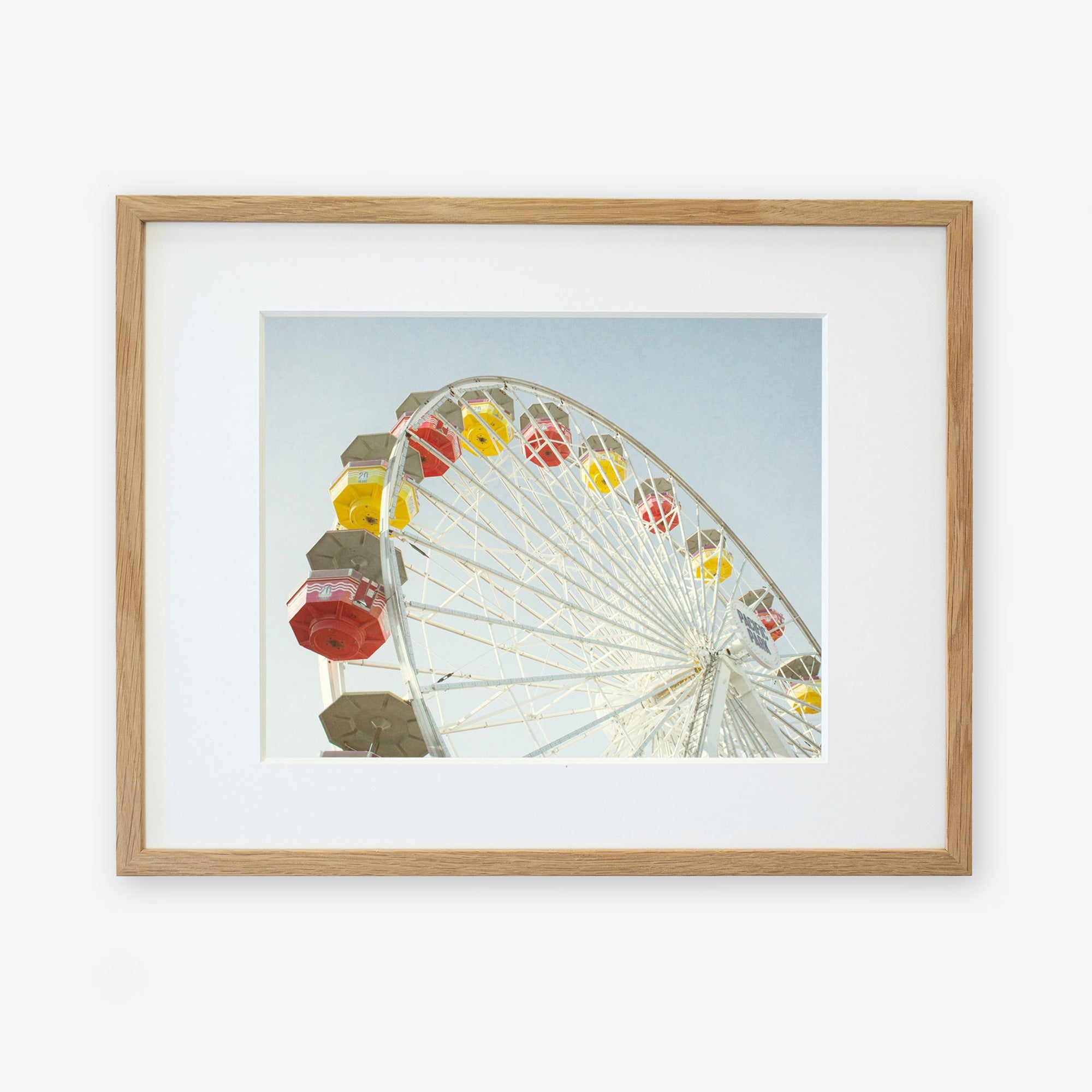 A framed photograph of a colorful Santa Monica Ferris Wheel Print, &#39;Ferris Above&#39;, at Santa Monica Pier, set against a clear sky, viewed from below. The frame is wooden and simple, emphasizing the vivid colors of the ride by Offley Green.