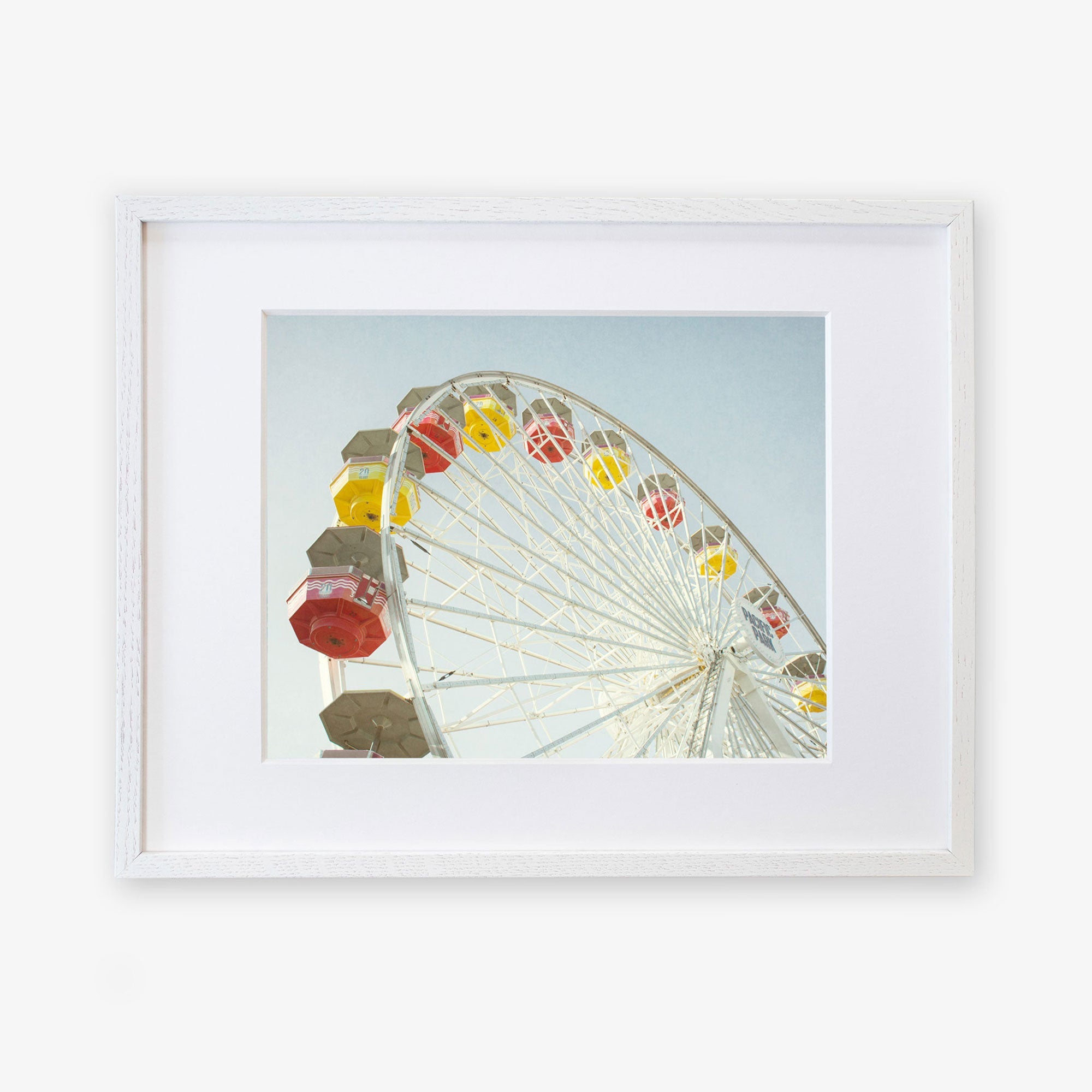 A framed photograph of a colorful Santa Monica Ferris Wheel Print, &#39;Ferris Above&#39; at Santa Monica Pier against a clear sky, displayed within a simple white frame on a white background by Offley Green.