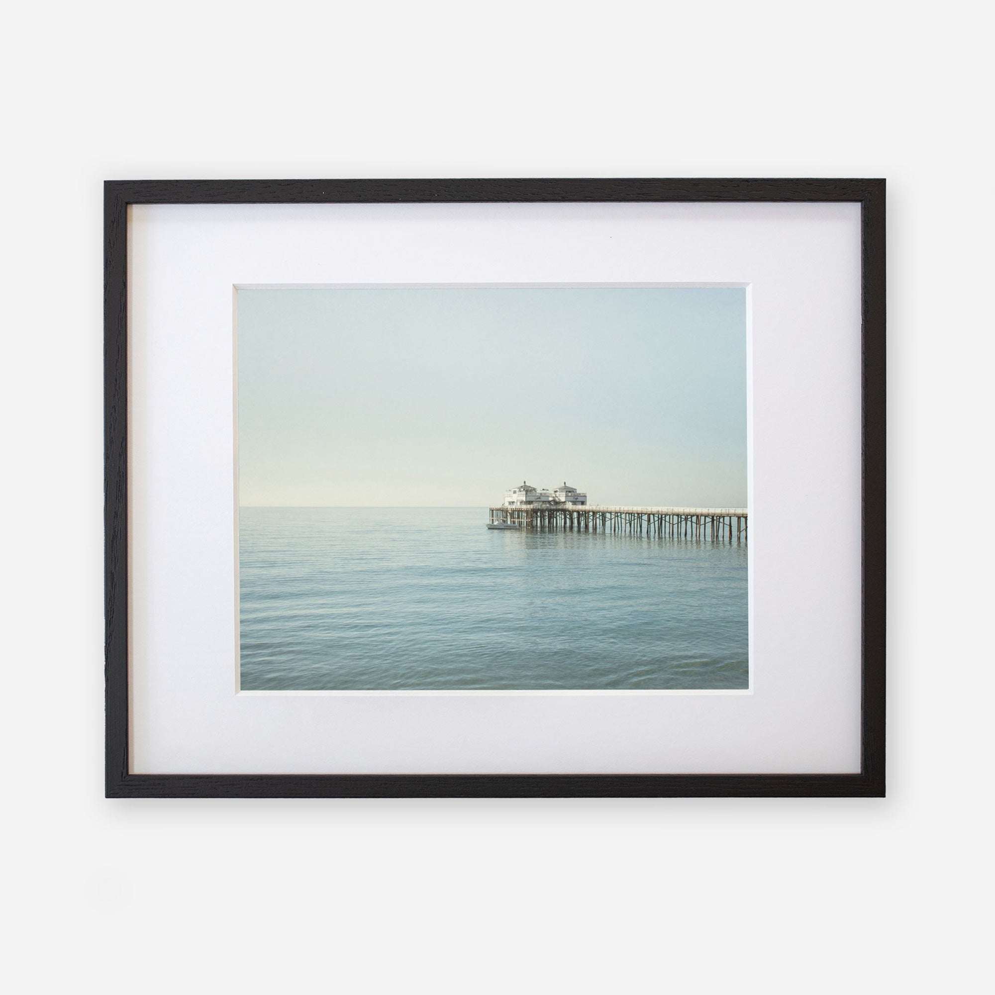 A framed photograph of a tranquil ocean scene featuring Malibu Pier extending into the water under a soft, misty sky. The frame is black with a white mat border, enhancing the image’s calm mood.
Product Name: Offley Green Coastal Print of Malibu Pier in California &#39;All Calm in Malibu&#39;