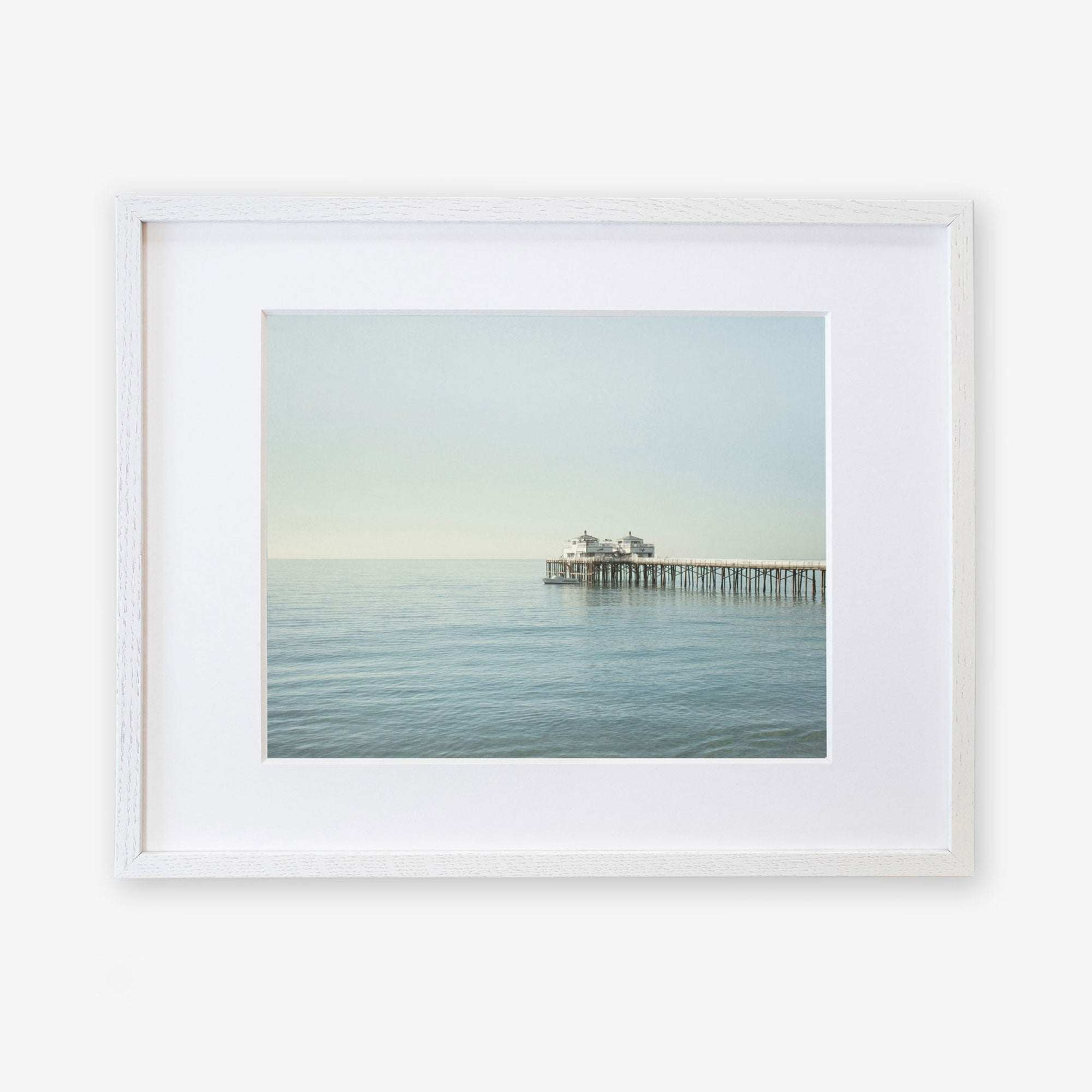 A framed photograph of a peaceful seascape with Malibu Pier extending into calm waters, displayed against a white background - Coastal Print of Malibu Pier in California &#39;All Calm in Malibu&#39; by Offley Green.