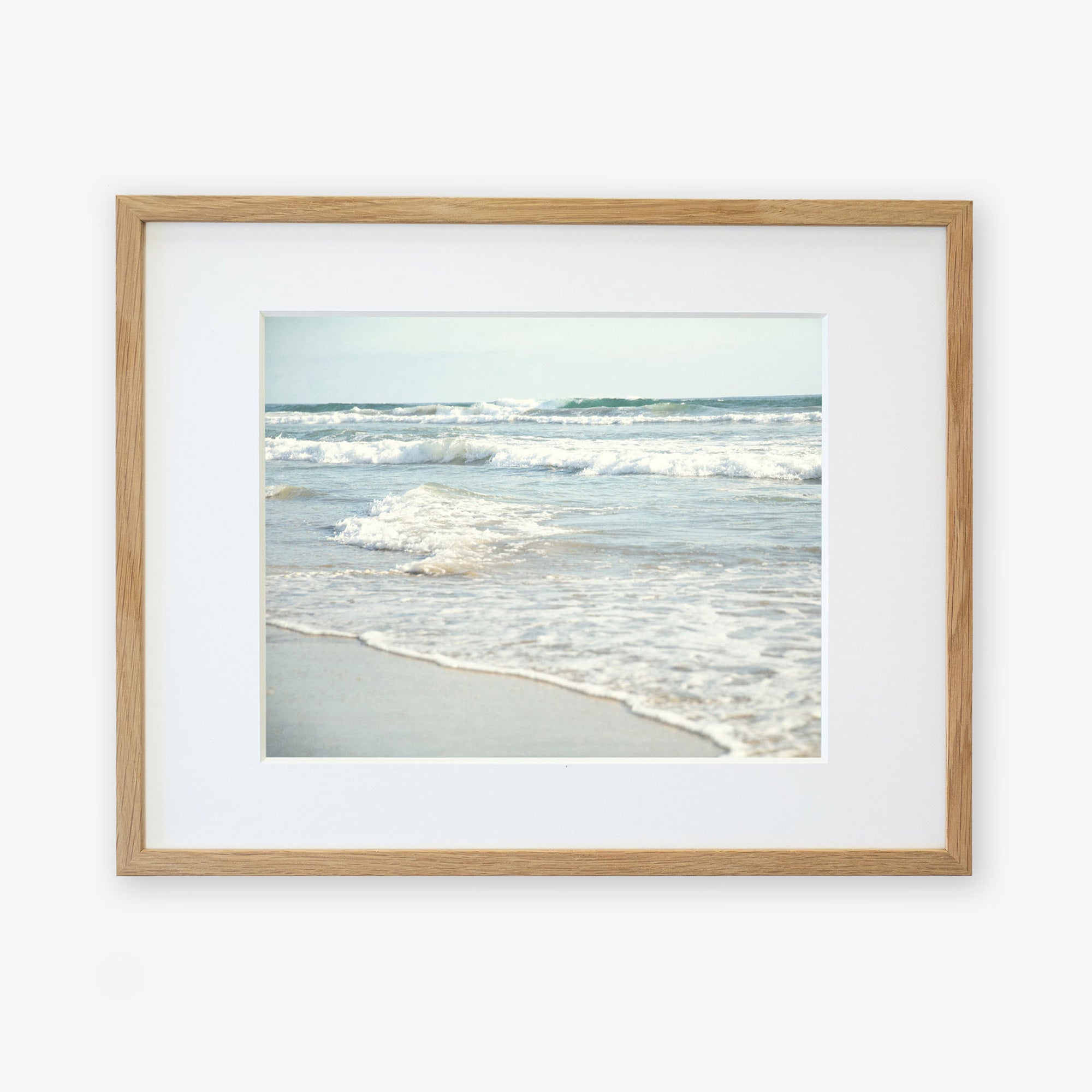 A framed photograph of a tranquil Carlsbad State Beach scene, with gentle waves lapping at the shore under a clear sky. The frame is simple and made of light wood, mounted on a white Offley Green Coastal Beach Print in California 'Surf and Sun'.