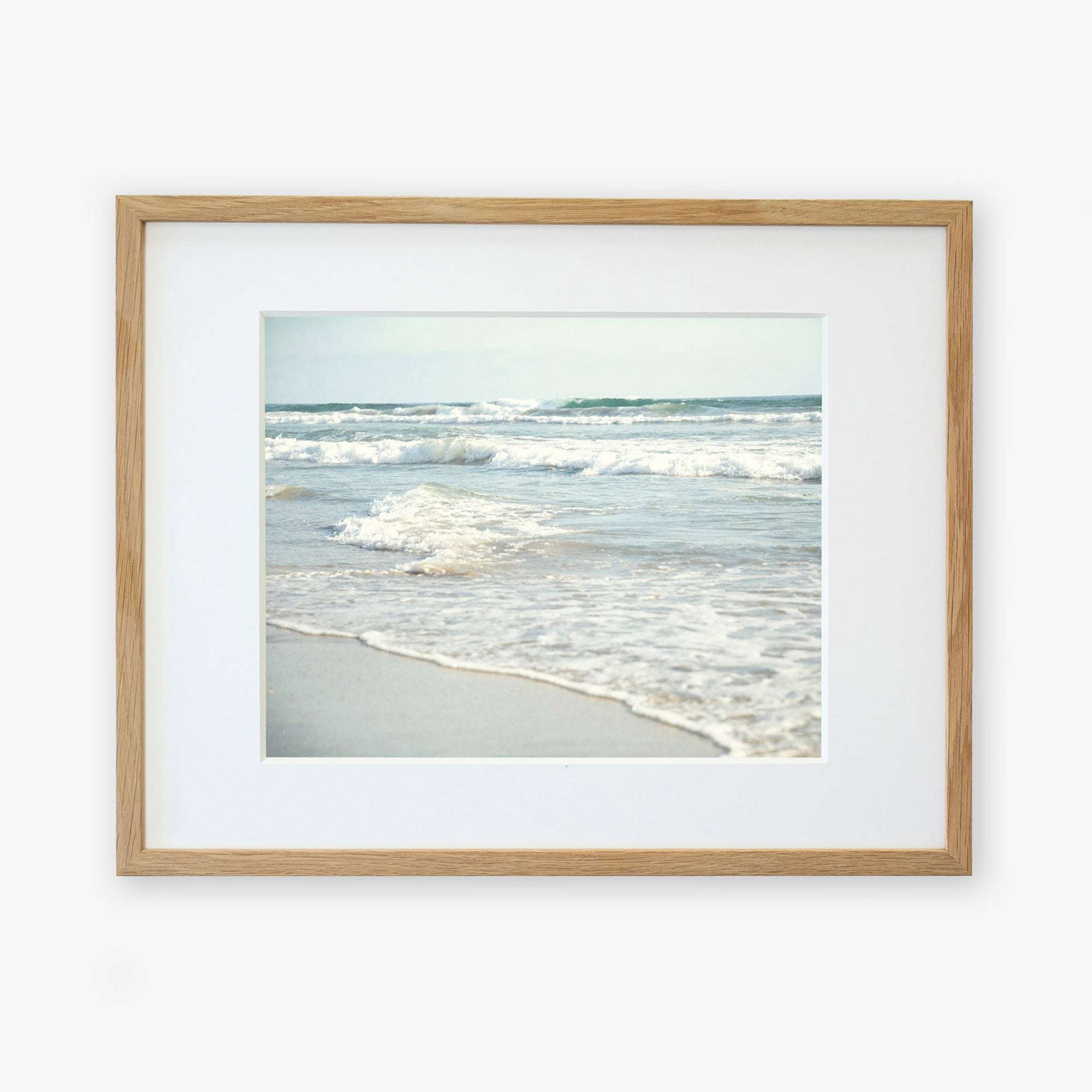 A framed photograph of a tranquil Carlsbad State Beach scene, with gentle waves lapping at the shore under a clear sky. The frame is simple and made of light wood, mounted on a white Offley Green Coastal Beach Print in California 'Surf and Sun'.