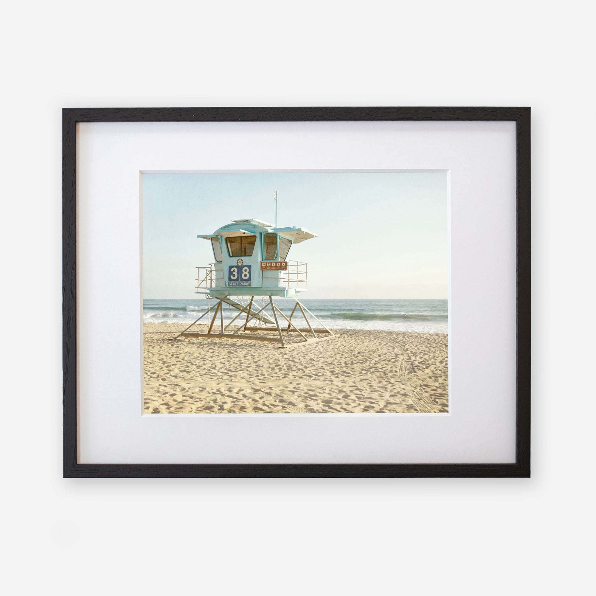 A framed photograph of a California Coastal Print, &#39;Carlsbad Lifeguard Tower&#39; on a sandy beach along the California coastline, with gentle waves in the background under a clear sky by Offley Green.
