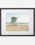 A framed photograph of a California Coastal Print, 'Carlsbad Lifeguard Tower' on a sandy beach along the California coastline, with gentle waves in the background under a clear sky by Offley Green.