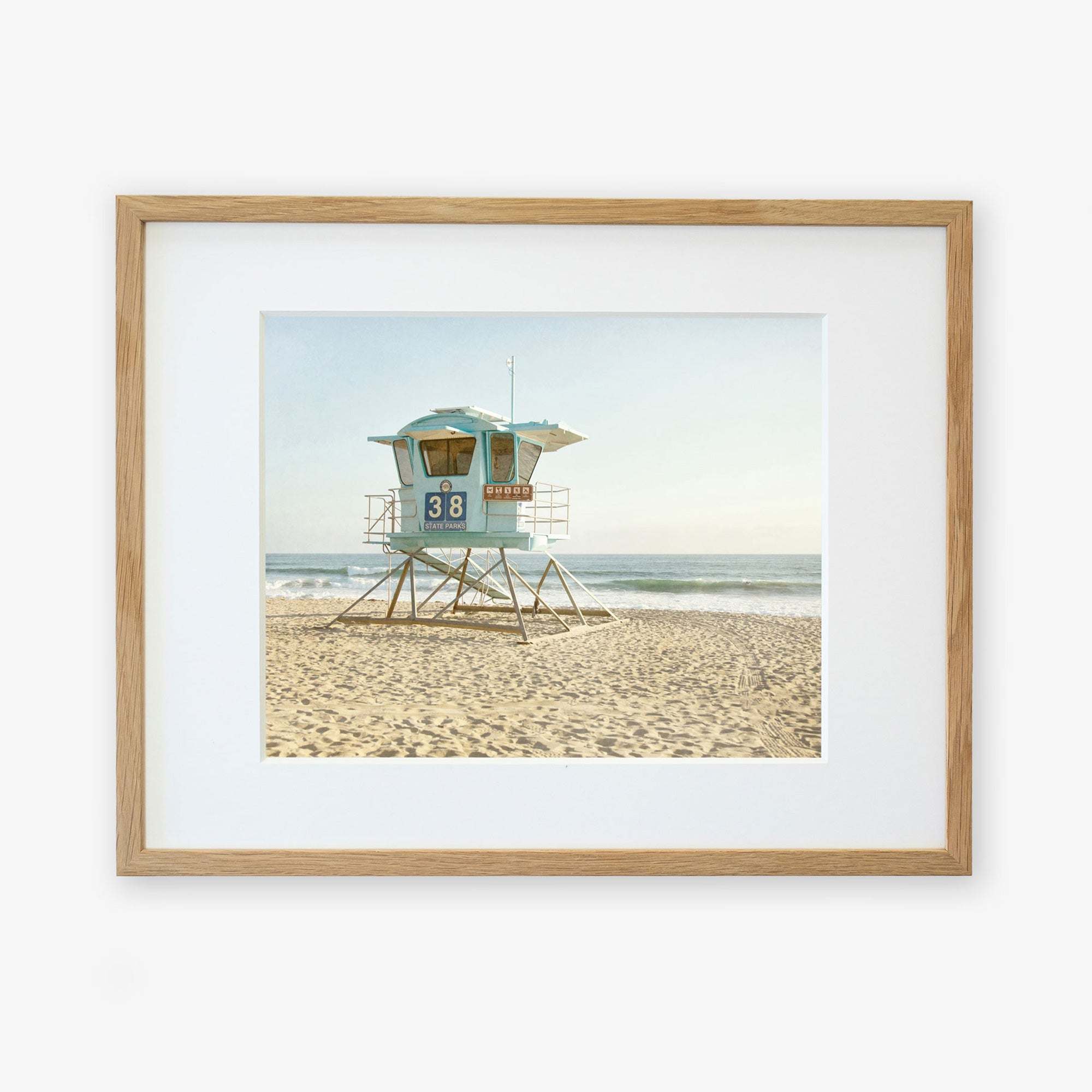 A framed photo of a lifeguard tower marked "38" standing on a sandy beach, printed on archival photographic paper, with serene ocean waves in the background under a clear sky - Offley Green's California Coastal Print, 'Carlsbad Lifeguard Tower'