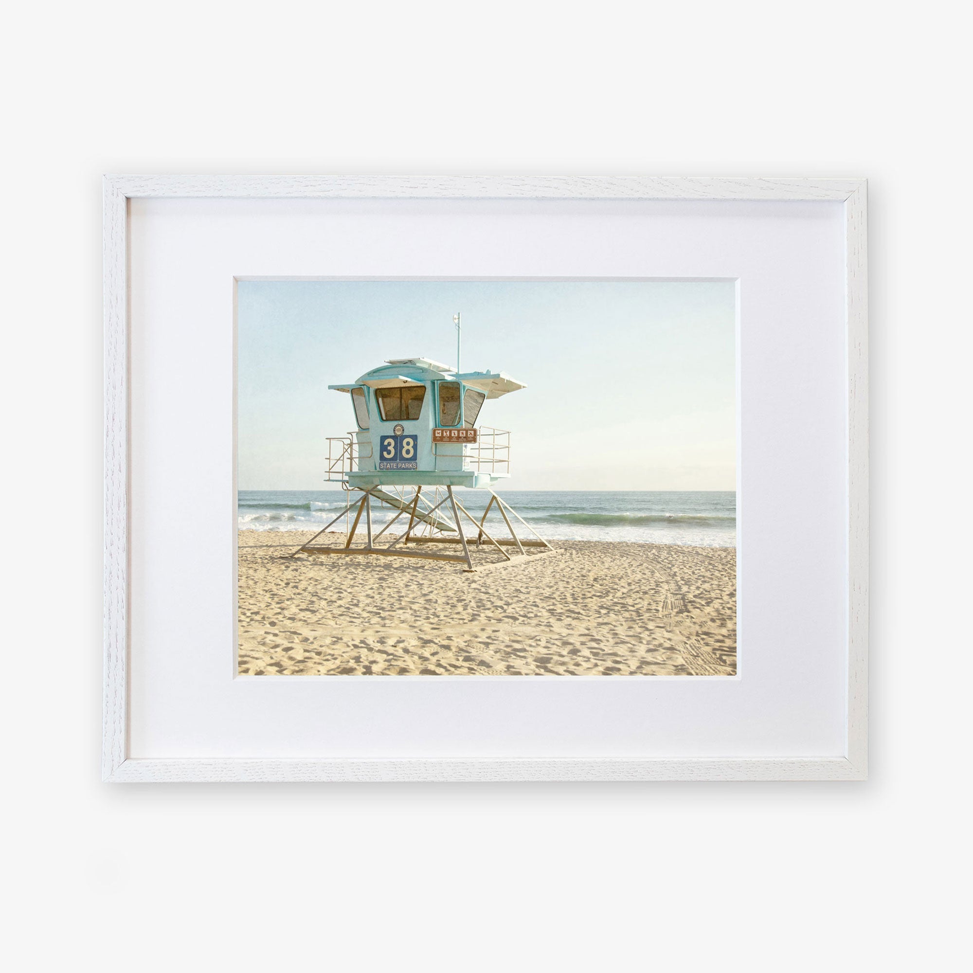 An unframed photograph of a lifeguard tower numbered 38 on a sandy beach, with the ocean in the background under a clear sky. The Offley Green 'California Coastal Print, Carlsbad Lifeguard Tower' exemplifies a serene beach setting.