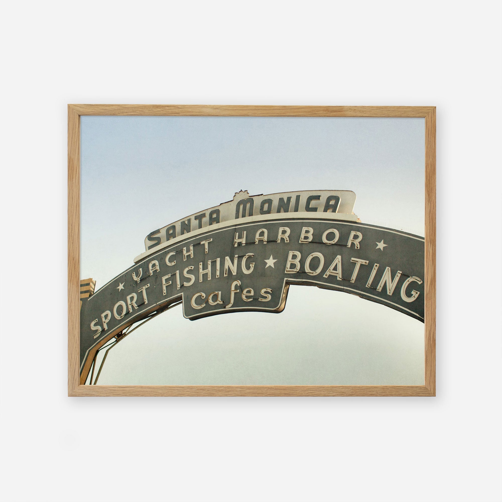 A framed archival photographic print of the iconic Santa Monica yacht harbor sign, featuring words like &quot;sport fishing,&quot; &quot;boating,&quot; and &quot;cafes&quot; on an arched metallic structure, against a plain Offley Green Los Angeles California Print, &#39;Santa Monica Pier Blues&#39;