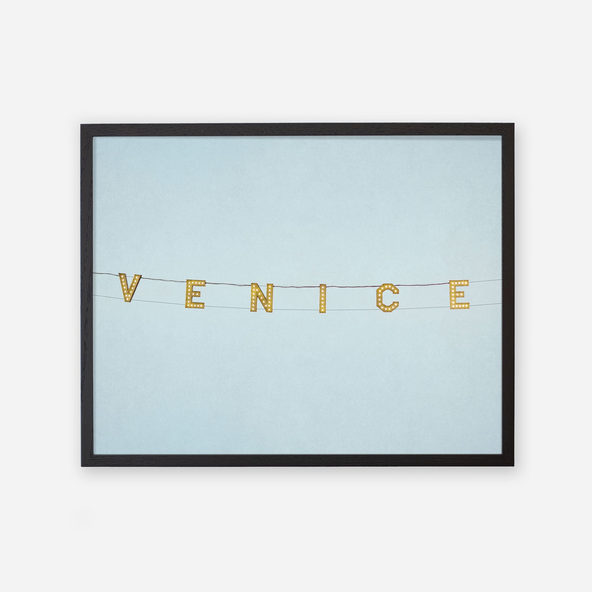 A framed picture of the &#39;Blue Venice&#39; Venice Beach Sign Print spelled out on a string of letter tiles against a plain light blue background, printed on archival photographic paper by Offley Green.