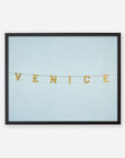 A framed picture of the 'Blue Venice' Venice Beach Sign Print spelled out on a string of letter tiles against a plain light blue background, printed on archival photographic paper by Offley Green.