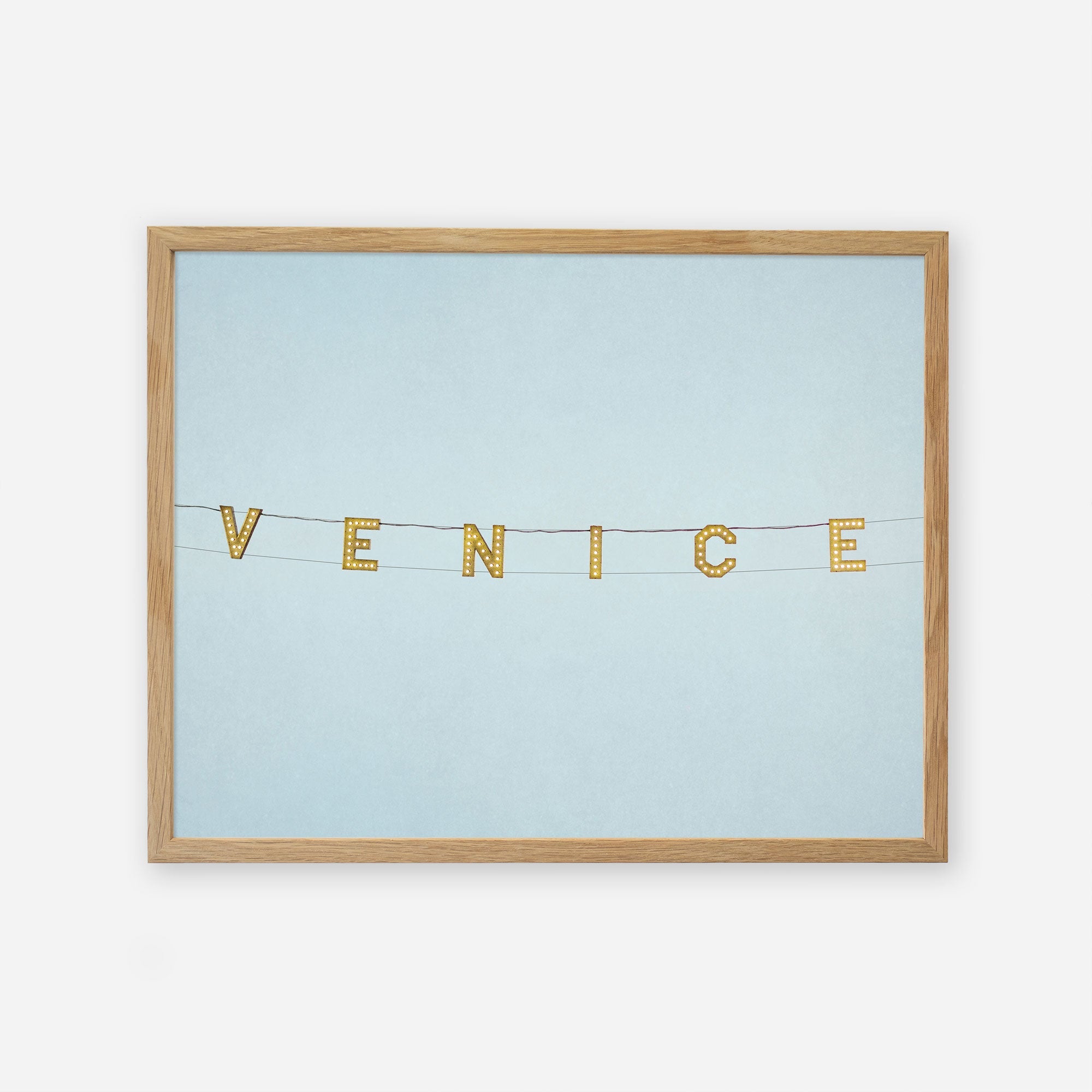 A light blue bulletin board displaying a decorative string with letters spelling &quot;Venice Beach Sign Print, &#39;Blue Venice&#39;&quot; in a horizontal arrangement, each letter on a separate, small flag by Offley Green.