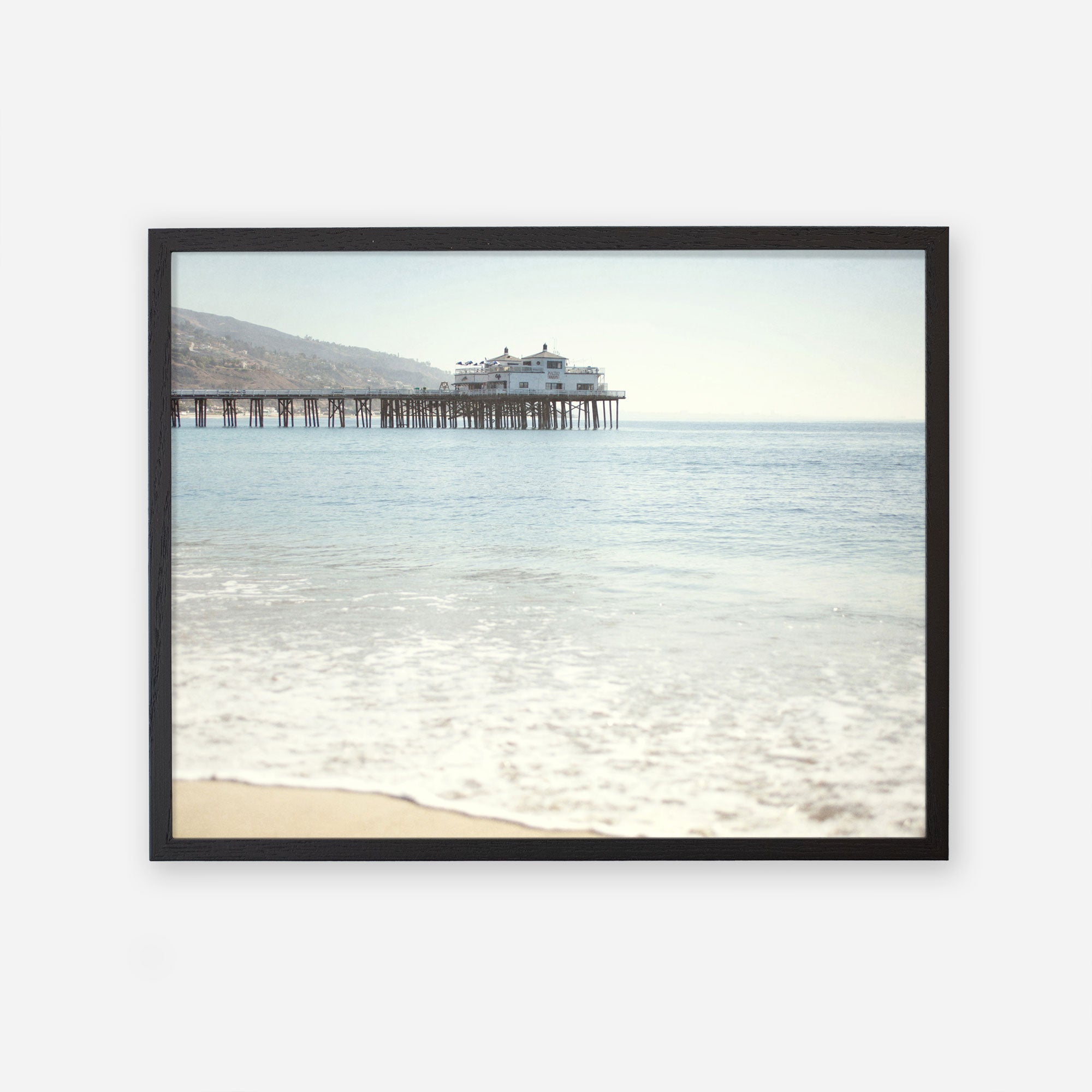 A framed photograph of a serene beach scene, showcasing gentle waves washing ashore with Malibu Pier extending into the ocean, topped by a small building under a clear sky. Offley Green&#39;s California Beach Print featuring &#39;Malibu Pier&#39;.