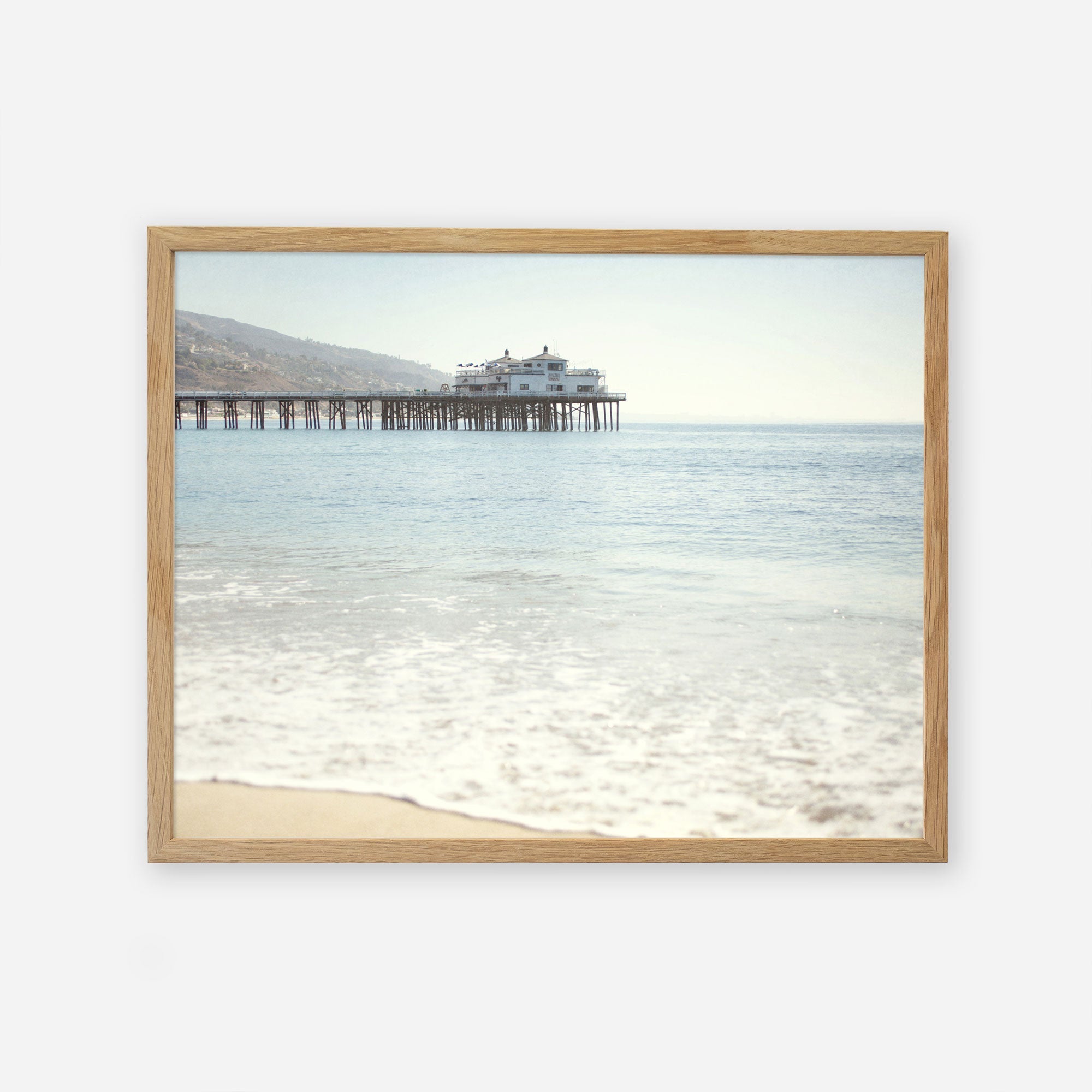 A framed photograph of a tranquil beach scene with gentle waves lapping at the shore and &#39;Malibu Pier&#39; extending into the ocean, supporting a large building under a clear sky. This is the Offley Green California Beach Print.