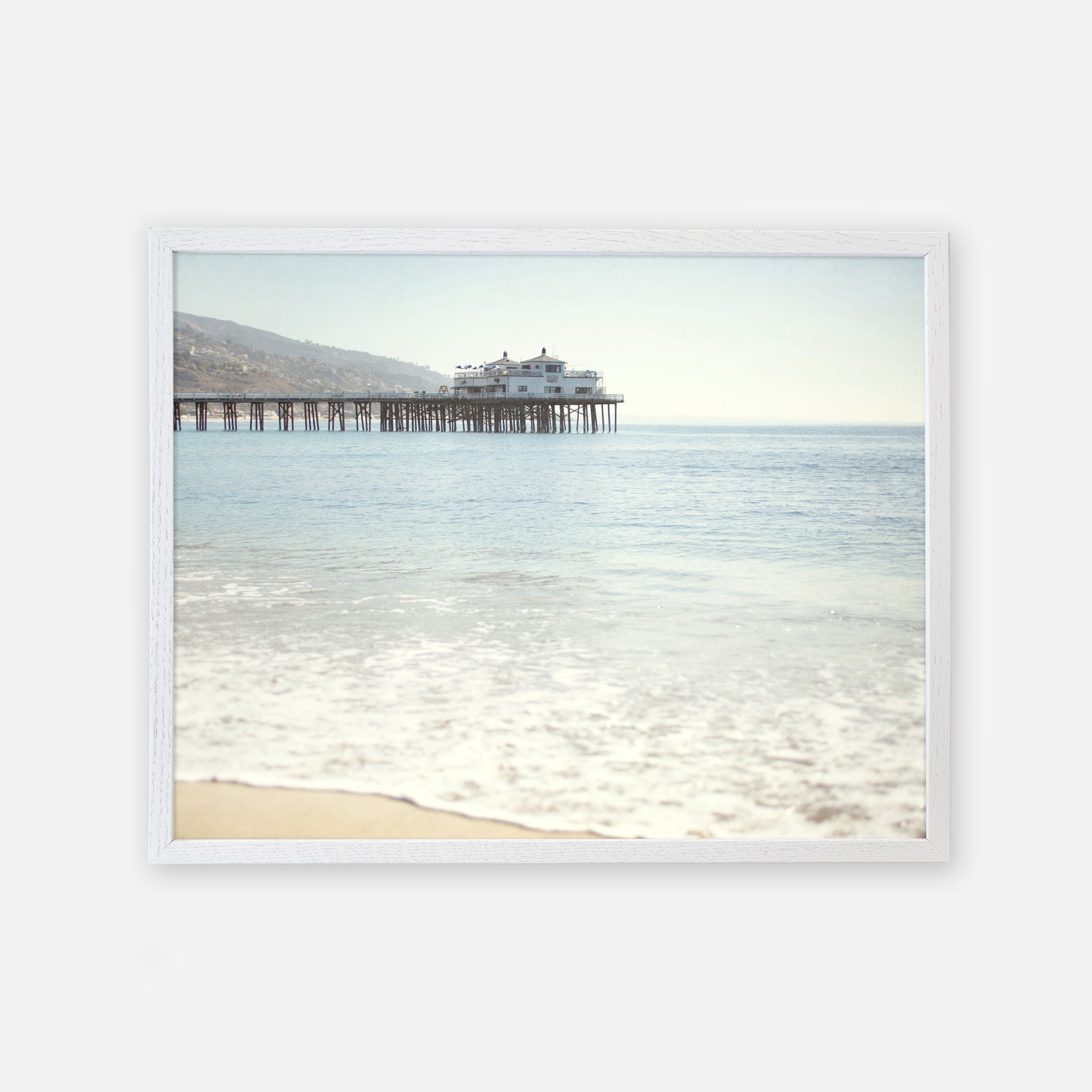 A serene beach scene with gentle waves lapping at the shore and a California Beach Print by Offley Green featuring Malibu Pier extending into the ocean, supporting a large building under a clear sky.