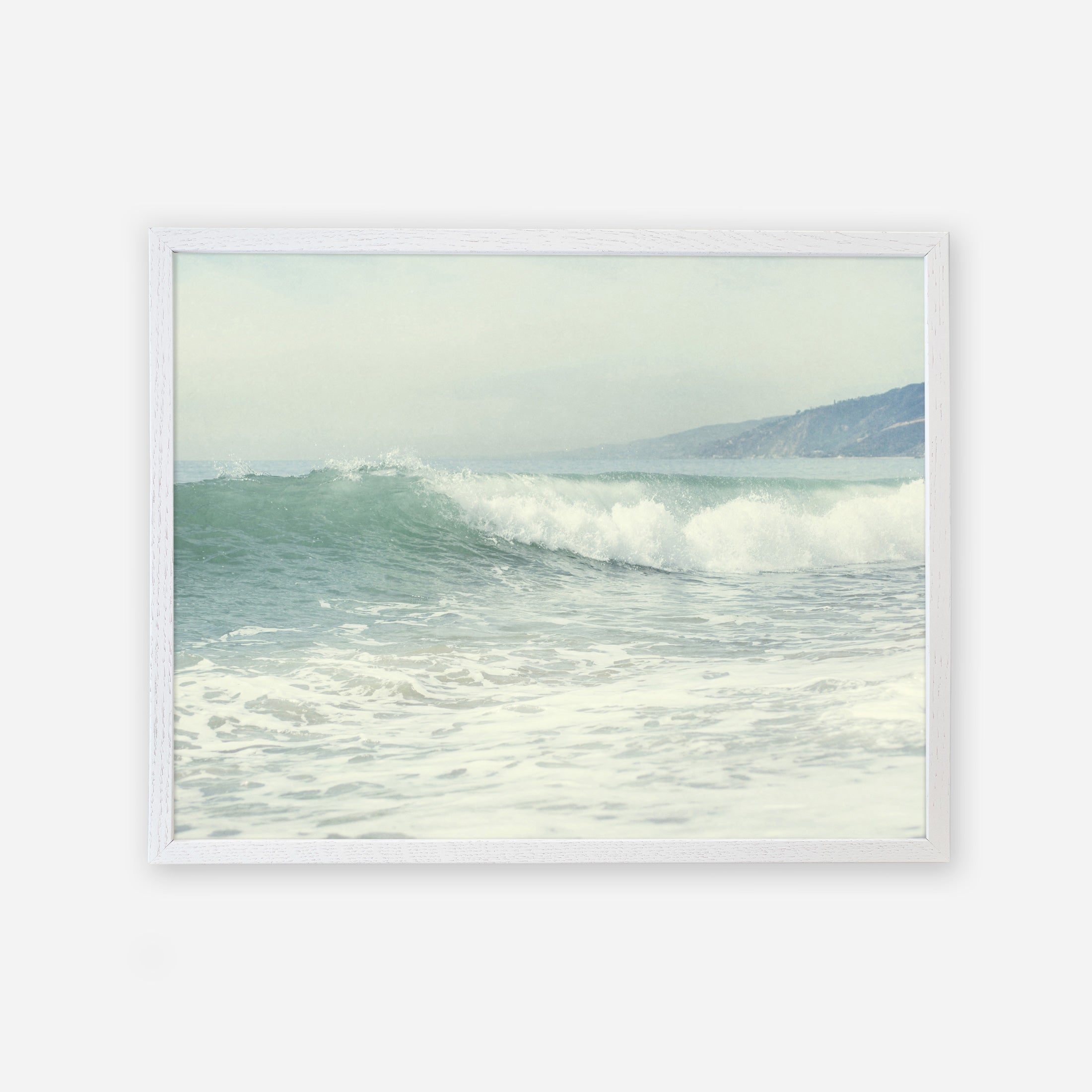 A framed illustration of gentle ocean waves rolling onto a Southern California beach, with hazy hills visible in the background under a soft, pale sky featuring the Offley Green Coastal Print of a Breaking Wave &#39;Breaking Surf&#39;.