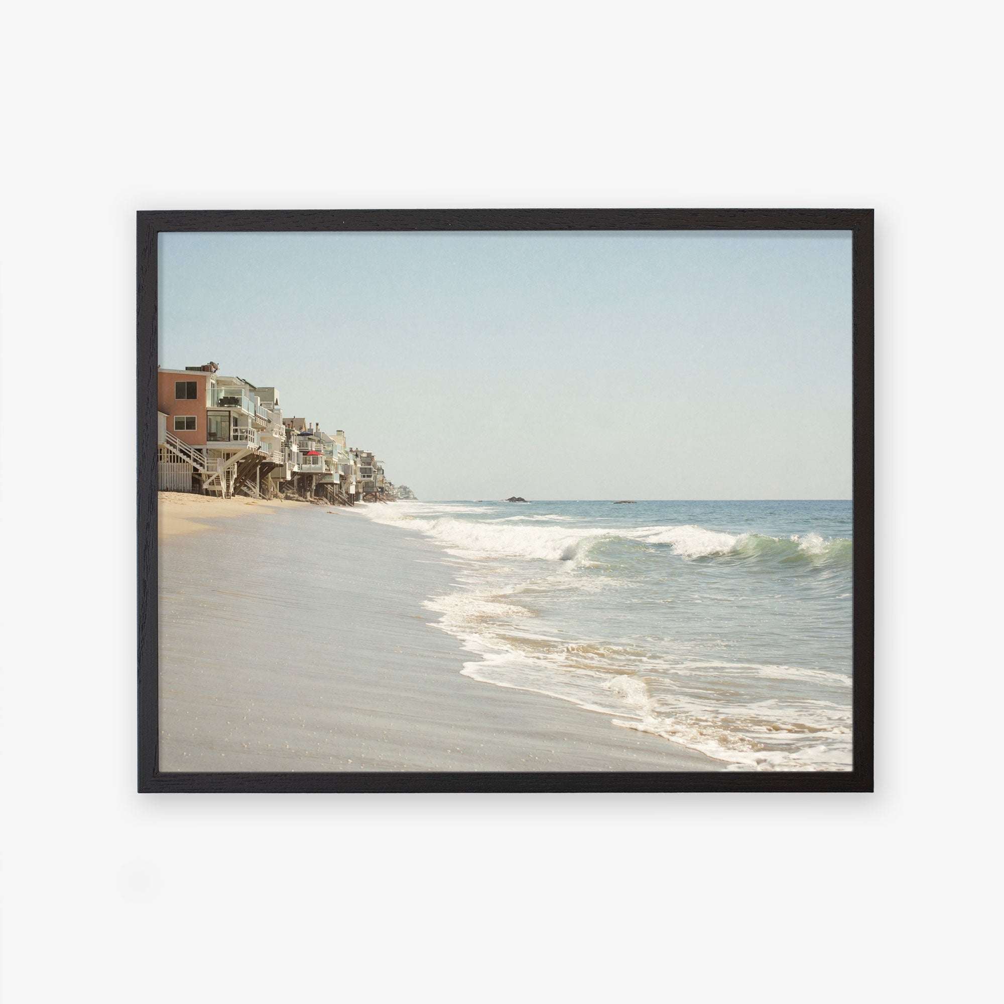 A framed wall art of a Malibu Beach House Print, &#39;Ocean View&#39; by Offley Green, showing beach houses perched along the edge of a sandy beach with gentle waves lapping the shore under a clear sky.