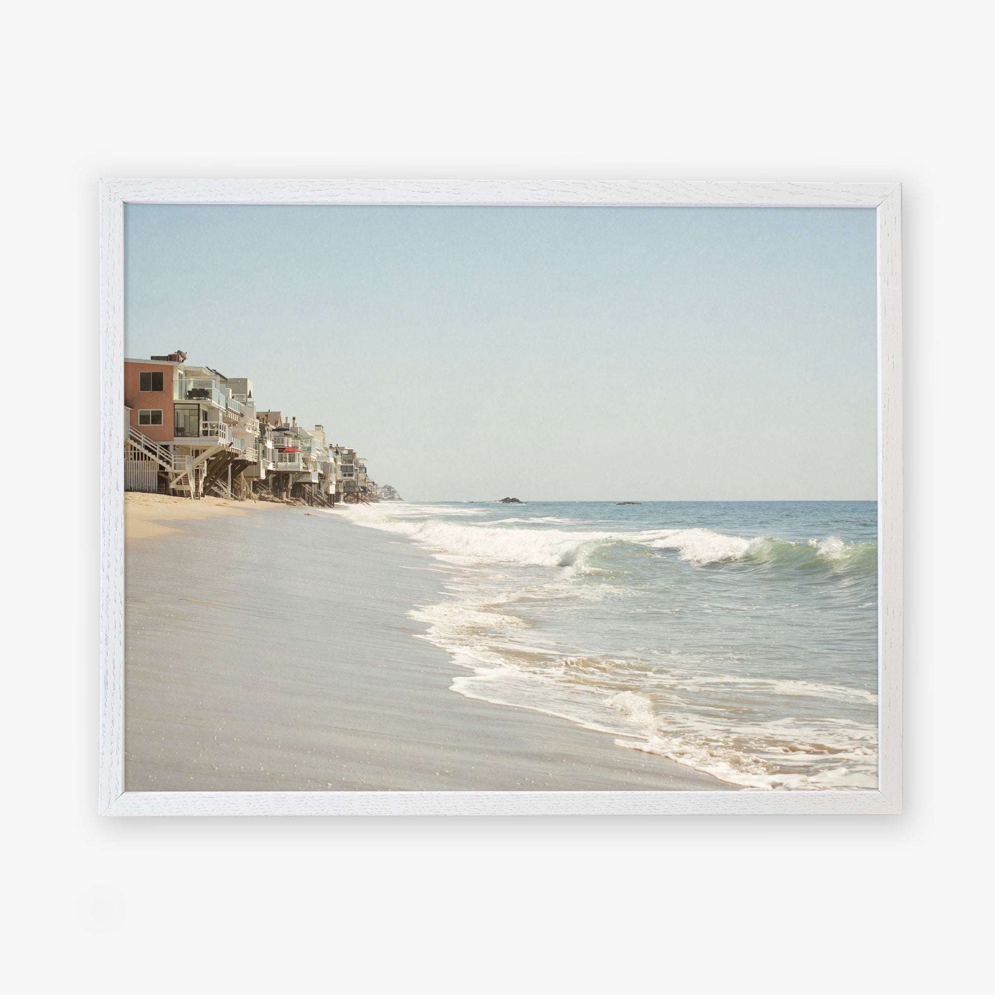 A framed wall art depicting a serene Malibu coastline with gentle waves, showcasing a row of houses built along the sandy shore under a clear sky. Offley Green&#39;s Malibu Beach House Print, &#39;Ocean View&#39;.