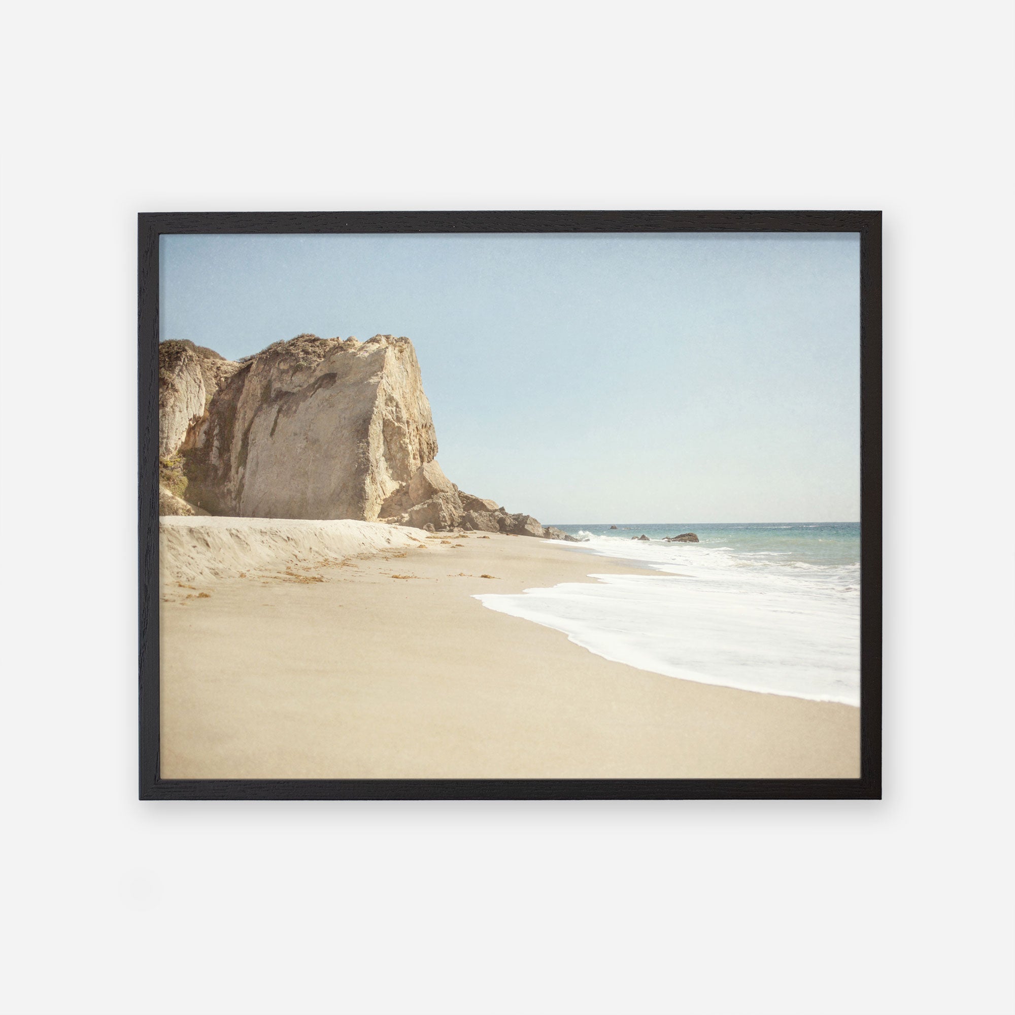 A framed photograph depicting a serene beach scene at Point Dume with a large cliff on the left, gentle waves washing onto the sandy shore, and a clear blue sky - Offley Green&#39;s California Malibu Print, &#39;Point Dume&#39;