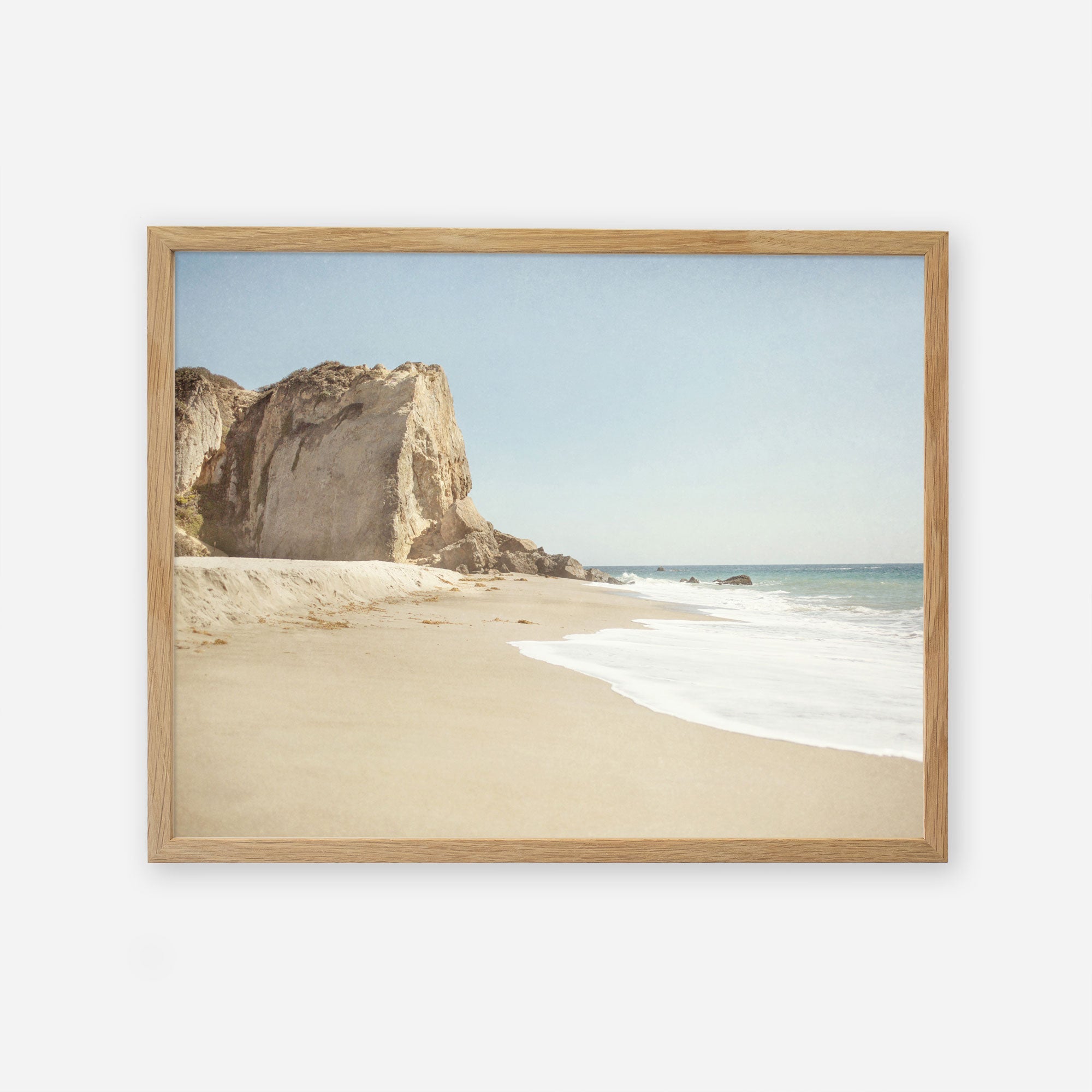 A framed painting of a serene beach scene at Point Dume, with a large cliff on the left, white sand, and gentle waves under a clear sky by Offley Green&#39;s California Malibu Print, &#39;Point Dume&#39;.