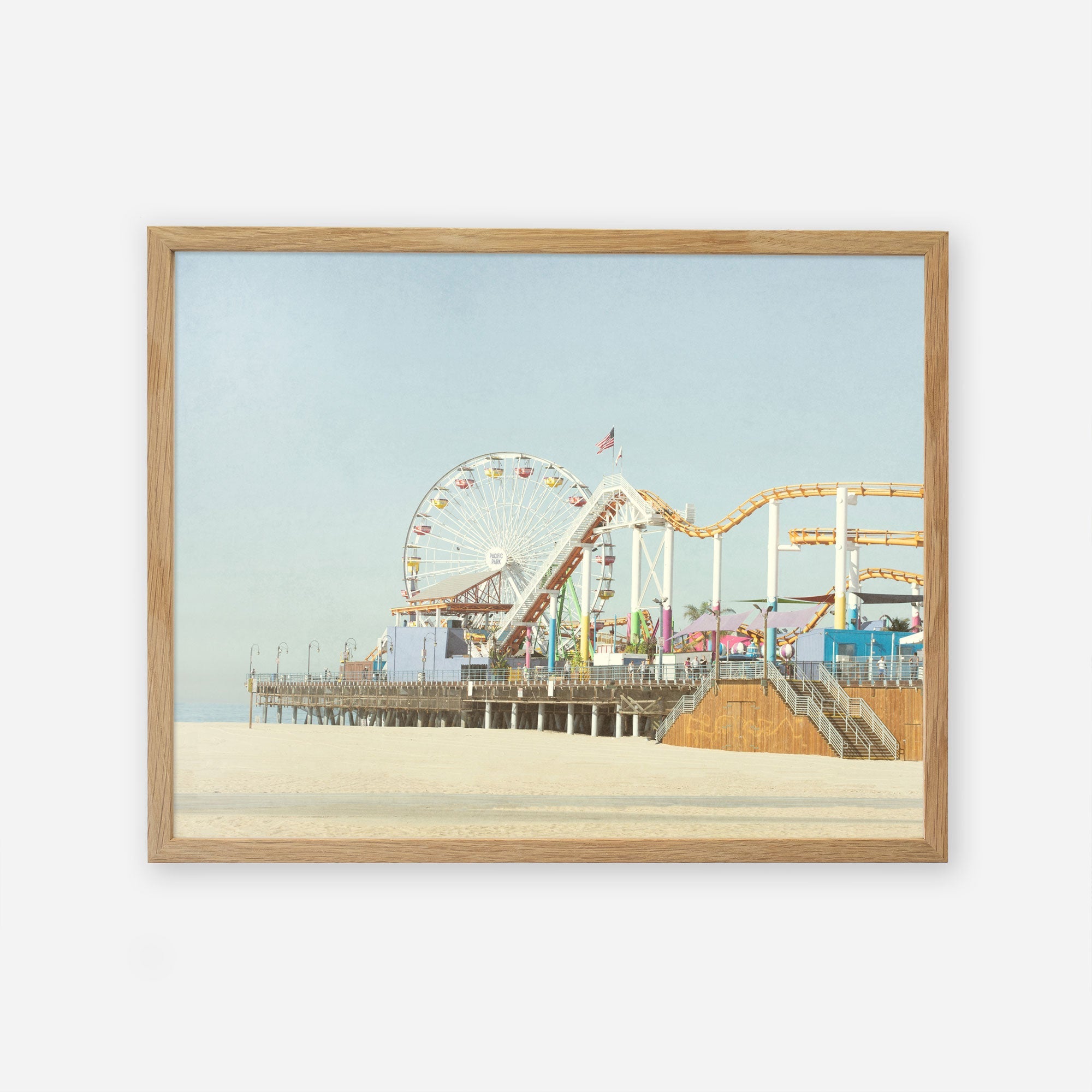 A framed painting of a lively Santa Monica Pier with a ferris wheel, roller coaster, and bustling visitors enjoying a sunny day, set against a clear blue sky - Offley Green&#39;s California Print, &#39;Santa Monica Pier&#39;.