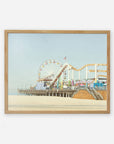 A framed painting of a lively Santa Monica Pier with a ferris wheel, roller coaster, and bustling visitors enjoying a sunny day, set against a clear blue sky - Offley Green's California Print, 'Santa Monica Pier'.