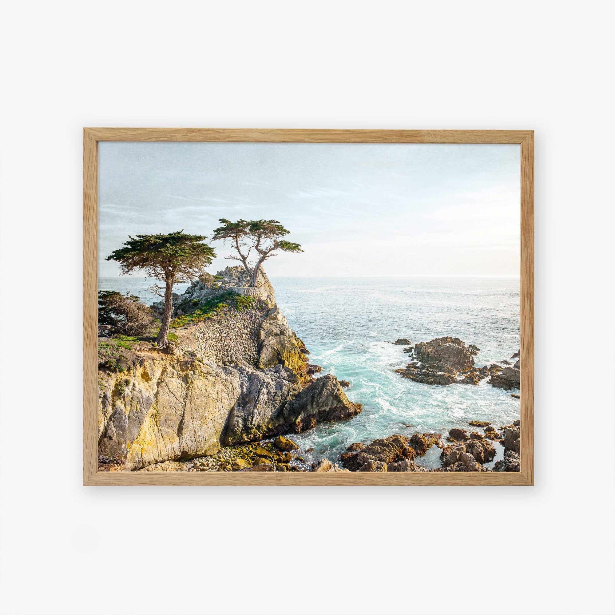 A framed painting depicting a scenic coastal landscape with rugged cliffs, a few windswept trees, and Offley Green&#39;s &#39;California Coastal Print, &#39;Lone Cypress&#39; photography under a clear sky.