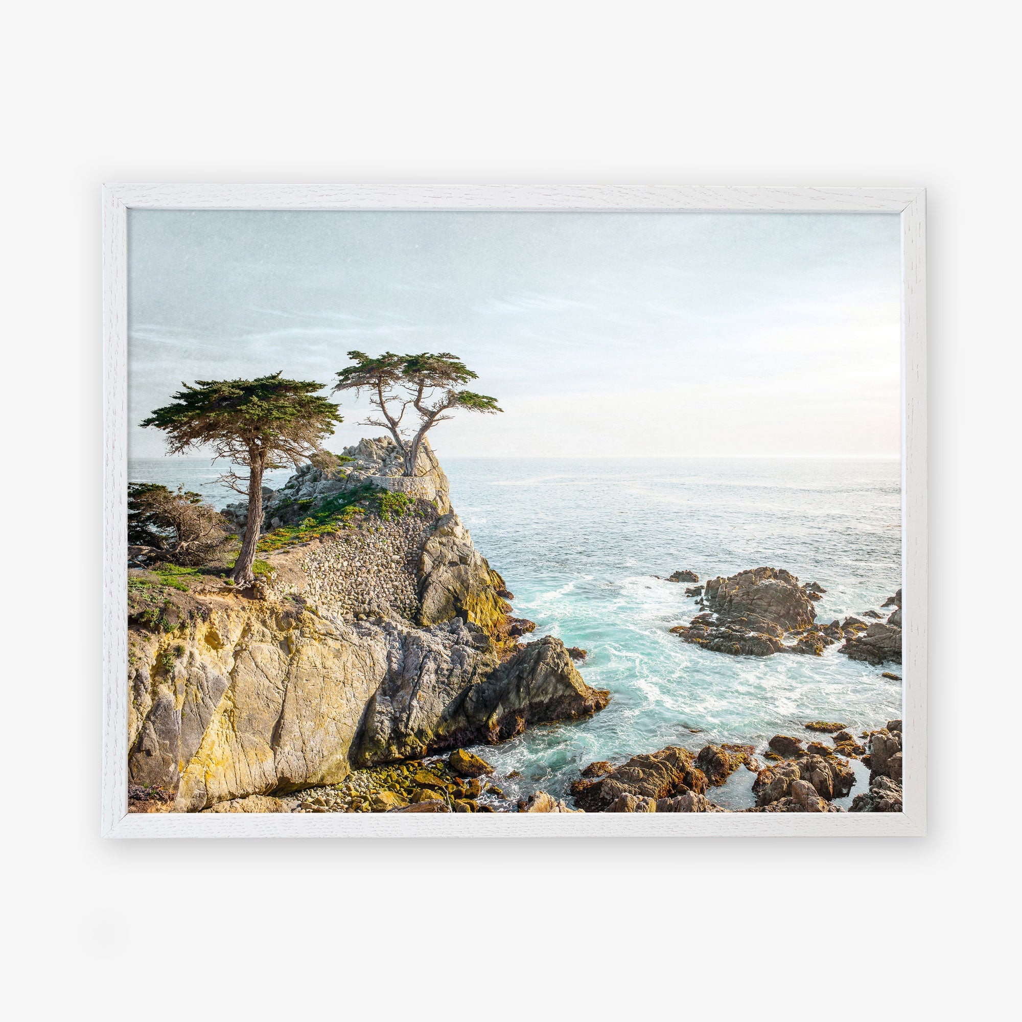 A framed painting of a rugged coastline with pine trees atop a cliff overlooking the sea at Pebble Beach, under a cloudy sky. The scene captures the serene and wild beauty of a coastal landscape. This is the Offley Green California Coastal Print, &#39;Lone Cypress&#39;.