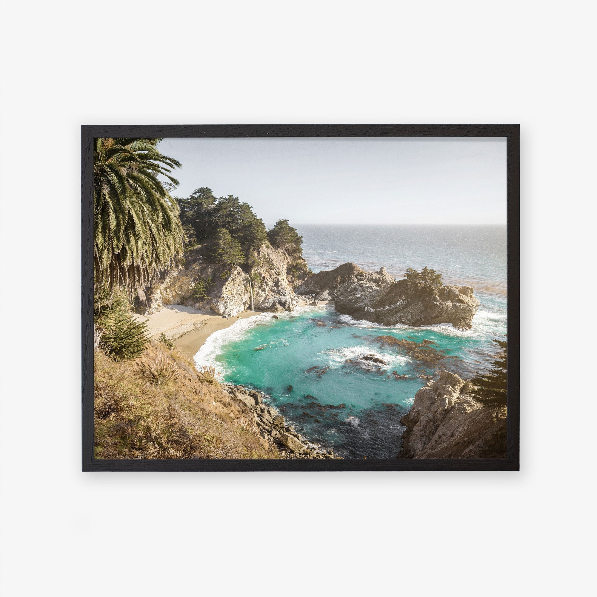 A framed photograph of a Offley Green Big Sur Coastal Print, &#39;Julia Pffeifer&#39; featuring rocky cliffs with a small sandy cove and turquoise waters, surrounded by lush greenery and tall palm trees.