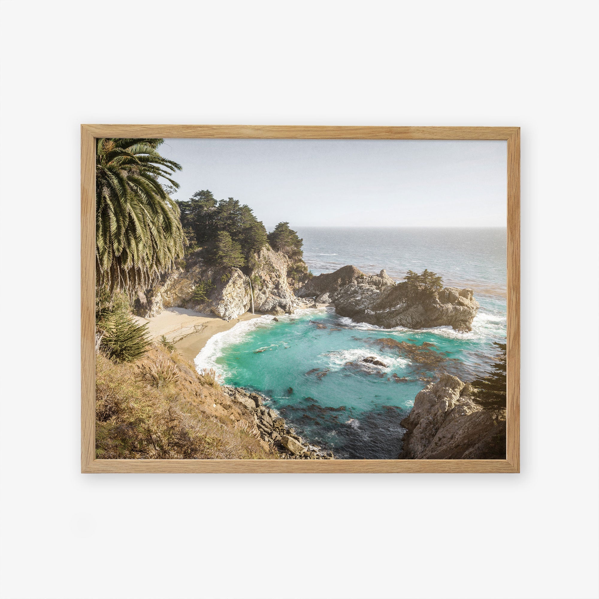 A framed scenic coastal landscape of Big Sur, showcasing a vibrant turquoise sea, rocky cliffs, and lush green trees under a clear blue sky. The product is the Offley Green Big Sur Coastal Print, &#39;Julia Pffeifer&#39;.