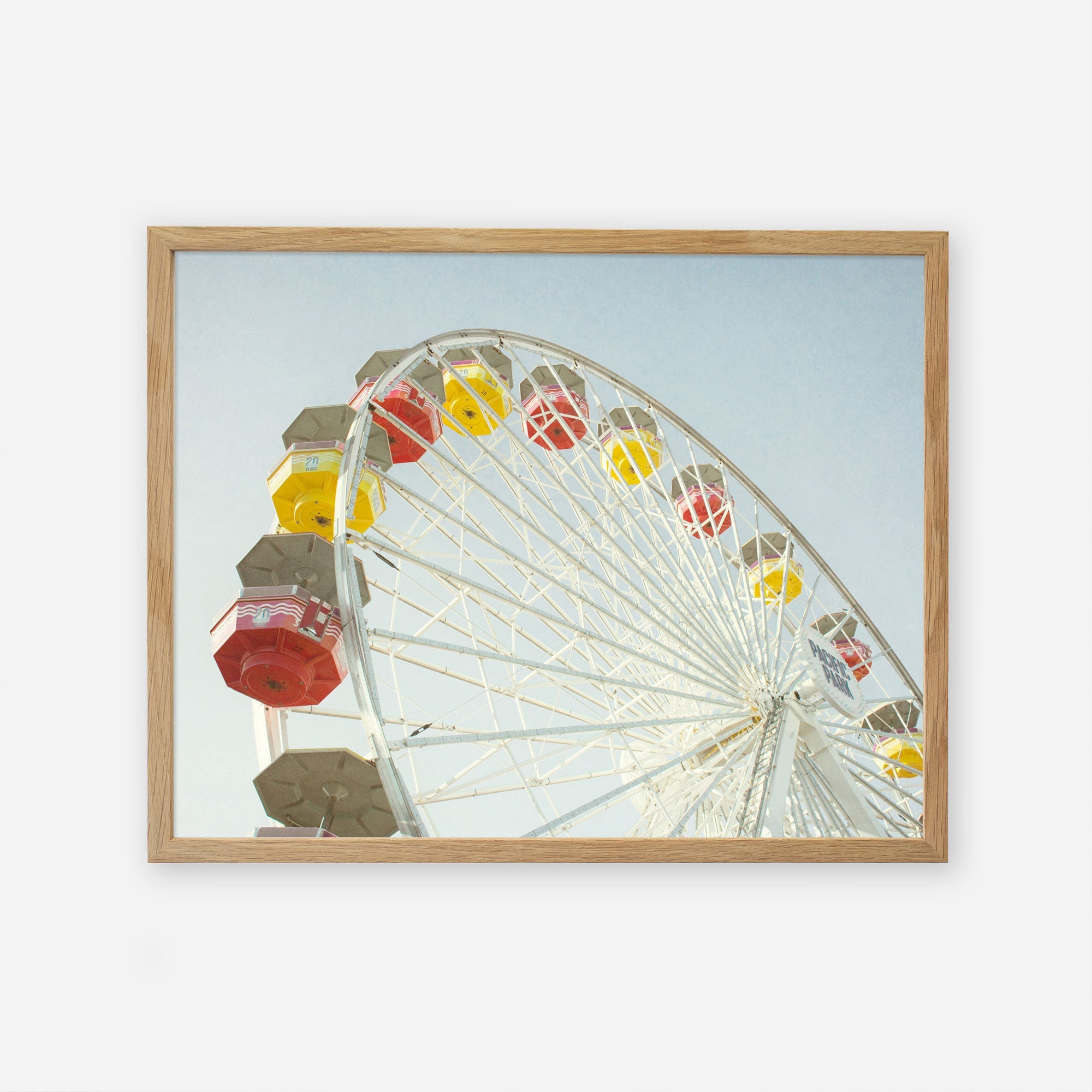 A framed photograph of a Santa Monica Ferris Wheel Print, &#39;Ferris Above&#39; with yellow, red, and gray cabins under a clear sky at Santa Monica Pier, centered within a simple wooden frame against a white background. (Offley Green)