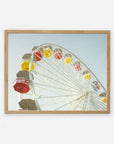 A framed photograph of a Santa Monica Ferris Wheel Print, 'Ferris Above' with yellow, red, and gray cabins under a clear sky at Santa Monica Pier, centered within a simple wooden frame against a white background. (Offley Green)