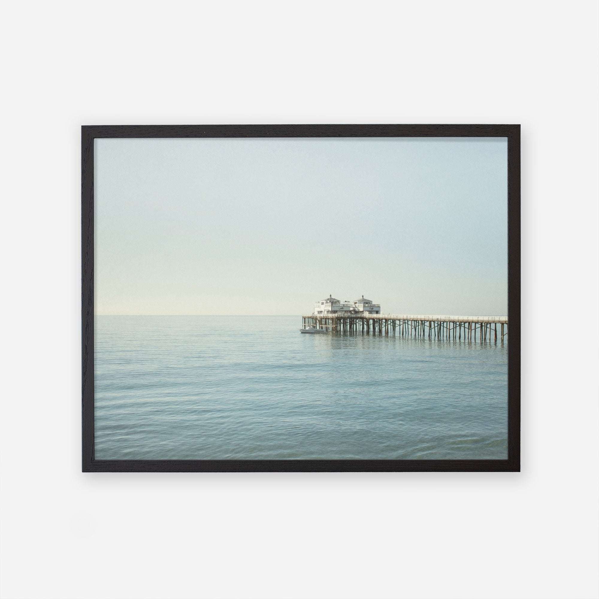 A framed photograph of a tranquil seascape featuring Malibu Pier with a small building extending into the calm ocean under a clear sky, Coastal Print of Malibu Pier in California &#39;All Calm in Malibu&#39; by Offley Green.