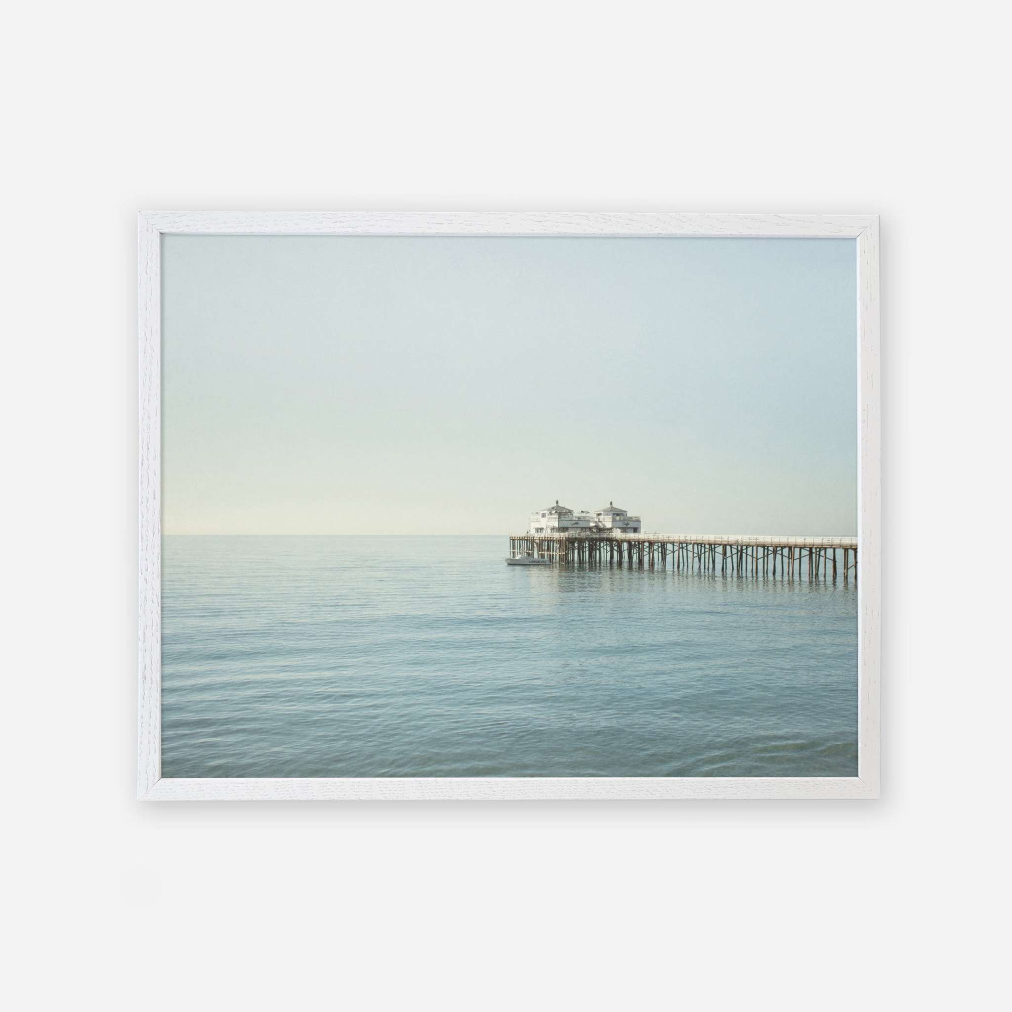 A framed photograph of a tranquil seascape with Malibu Pier extending into the calm sea under a soft, pale sky. The image conveys a serene, timeless atmosphere featuring the Coastal Print of Malibu Pier in California &#39;All Calm in Malibu&#39; by Offley Green.