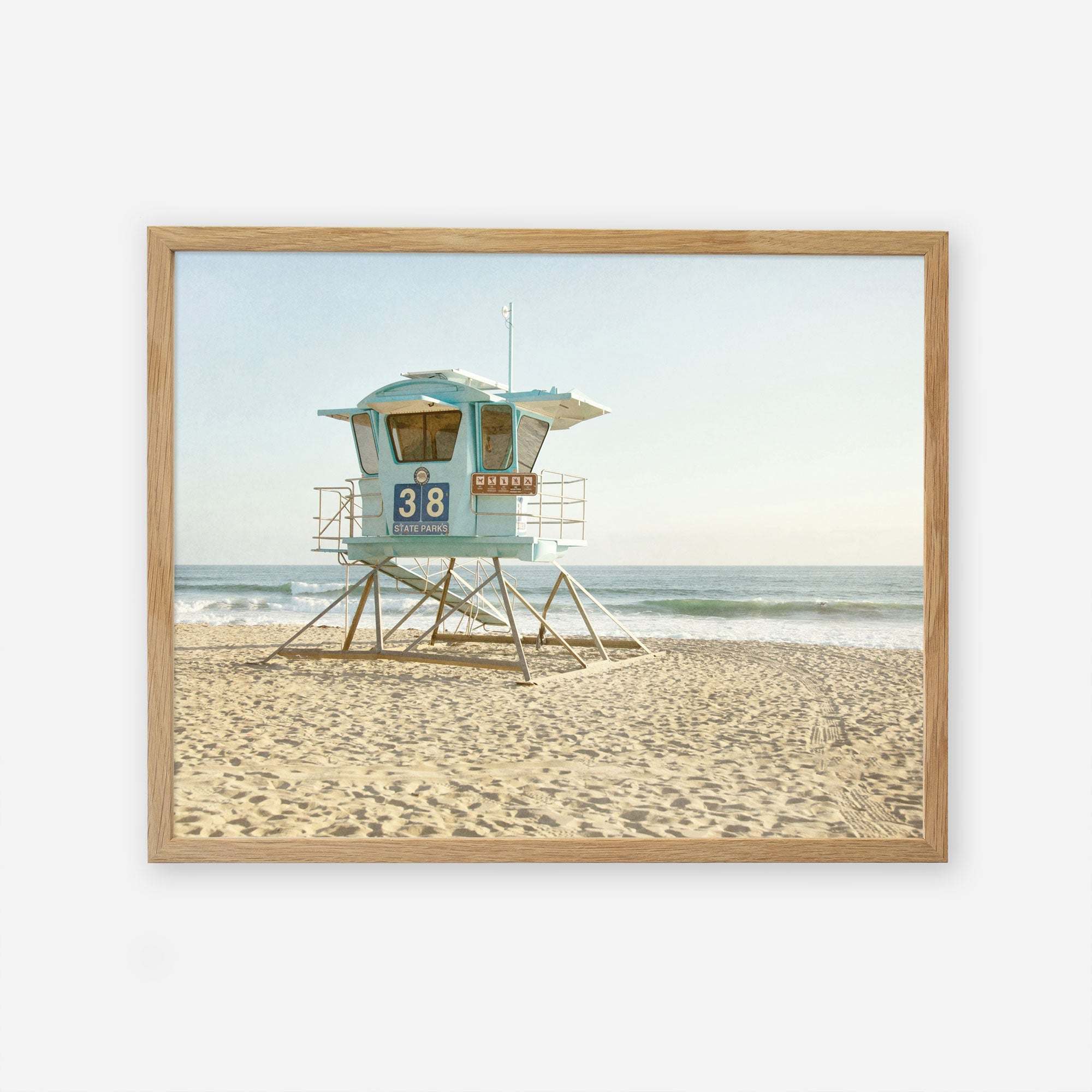 A framed painting of a California Coastal Print, &#39;Carlsbad Lifeguard Tower&#39; numbered 38 on a sandy beach with gentle waves in the background, under a clear sky. The tower features turquoise and white hues and captures the essence of California by Offley Green.