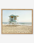 A framed painting of a California Coastal Print, 'Carlsbad Lifeguard Tower' numbered 38 on a sandy beach with gentle waves in the background, under a clear sky. The tower features turquoise and white hues and captures the essence of California by Offley Green.