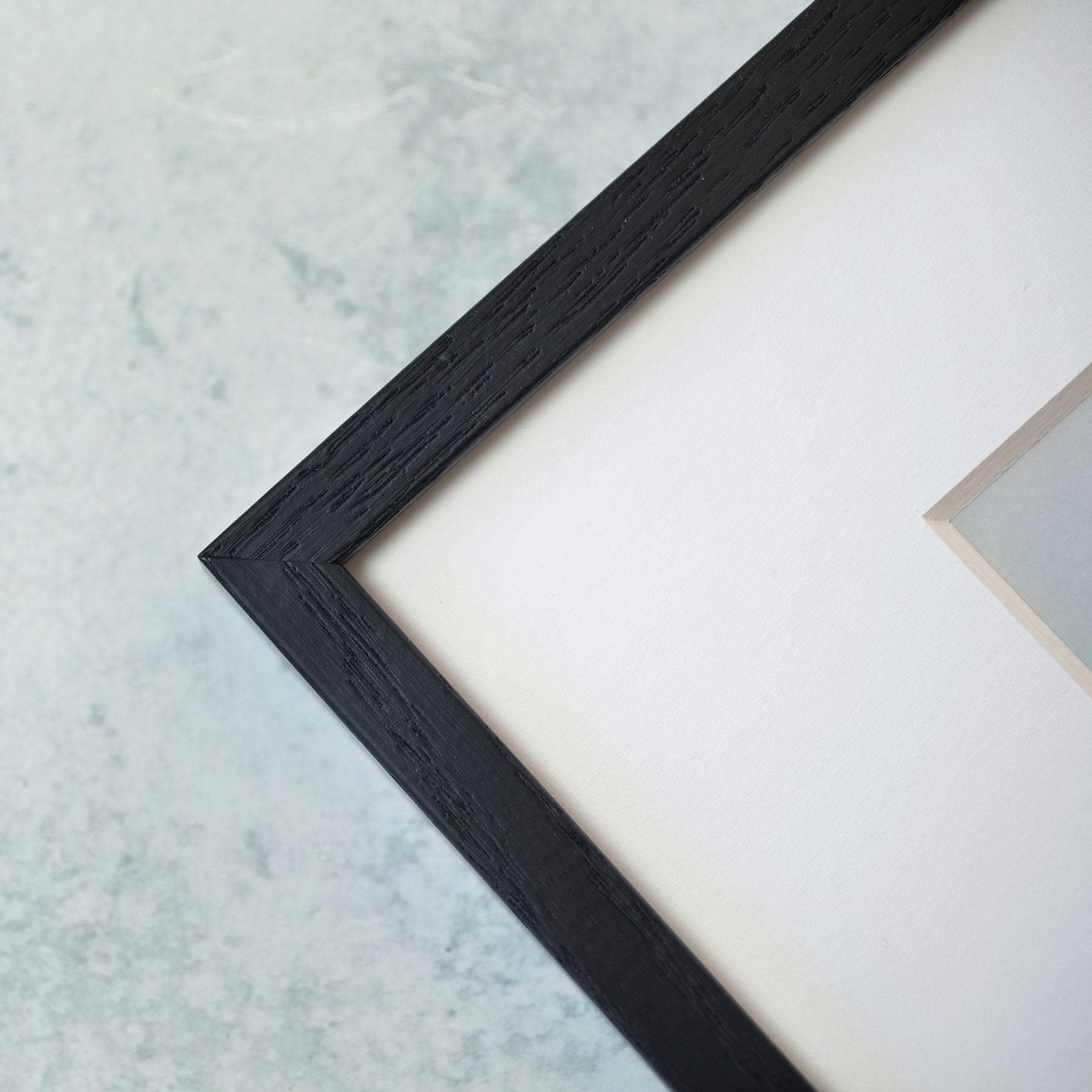 Close-up of a corner of a black textured &#39;Malibu Pier&#39; picture frame on a textured light blue background, partially enclosing an off-white canvas with coastal decor.