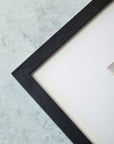 Close-up of a corner of a black textured 'Malibu Pier' picture frame on a textured light blue background, partially enclosing an off-white canvas with coastal decor.