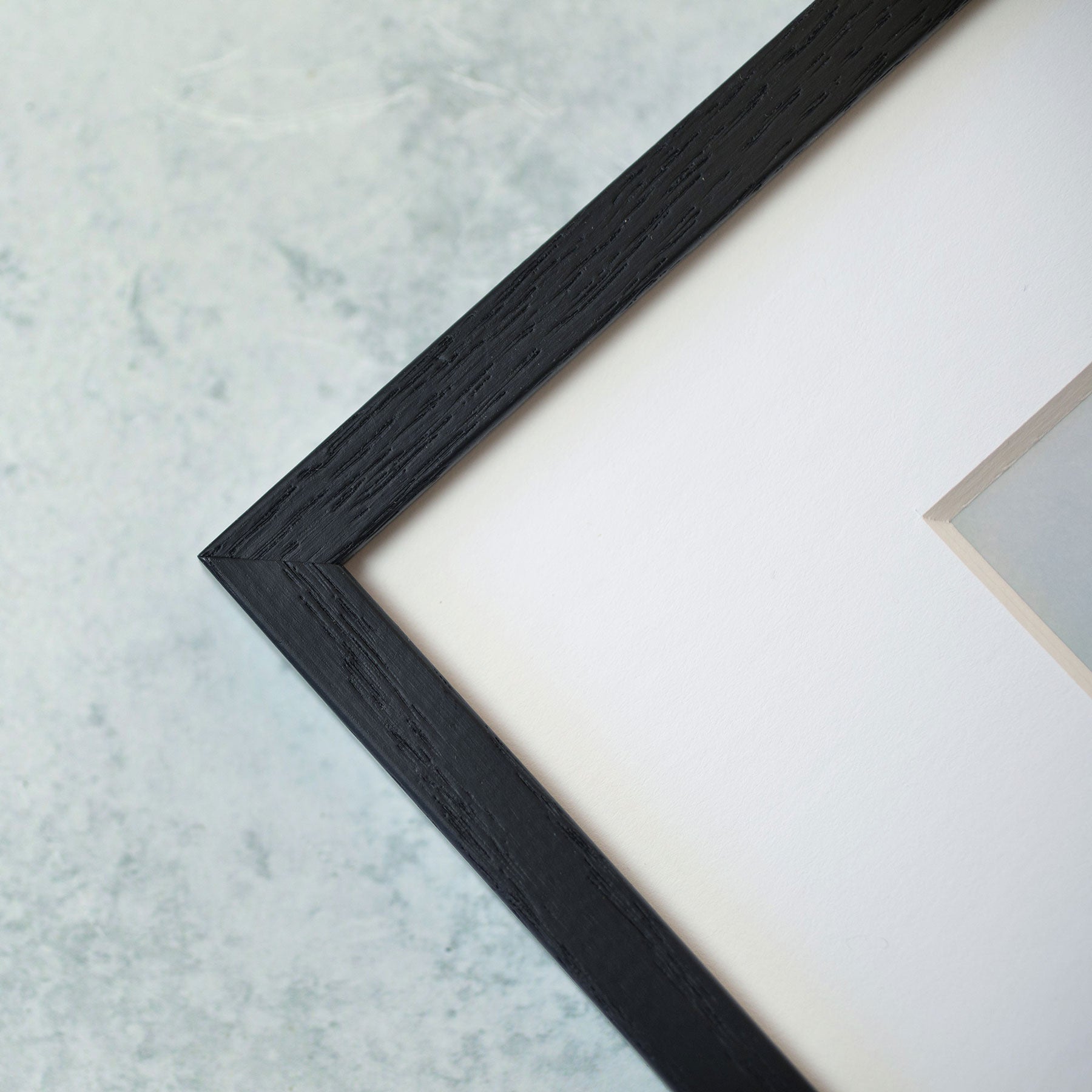 Close-up of the corner of a black wooden picture frame against a light, marble-like background. The frame is rectangular, showcasing an image of Seascape Coastal Art, &#39;Surf and Rocks&#39; by Offley Green printed on archival photographic paper with a moody seascape partially visible within the white-bordered picture.