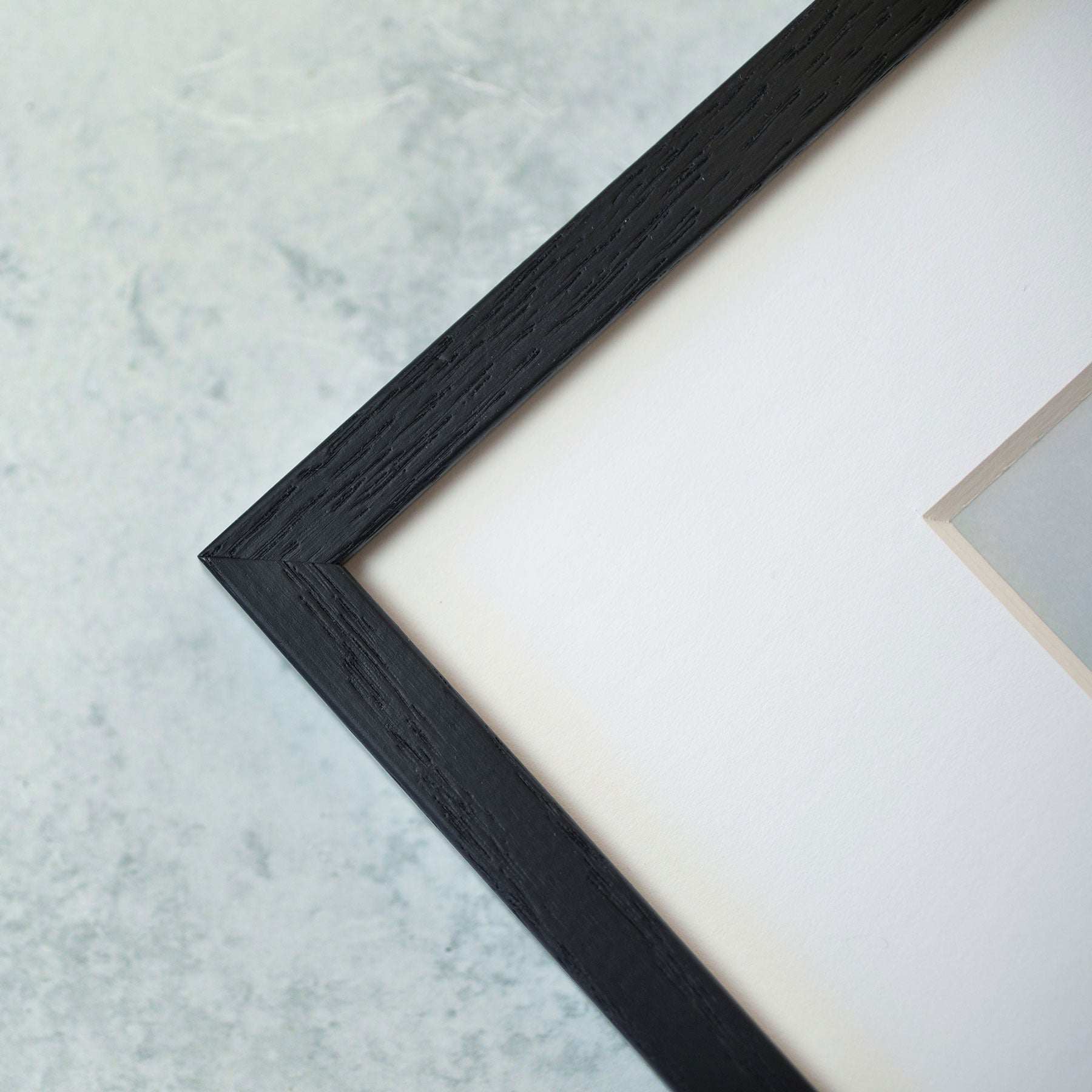 Close-up of a corner of a black textured Offley Green picture frame with a white border and a glimpse of a pale artwork on archival photographic paper against a light gray background.