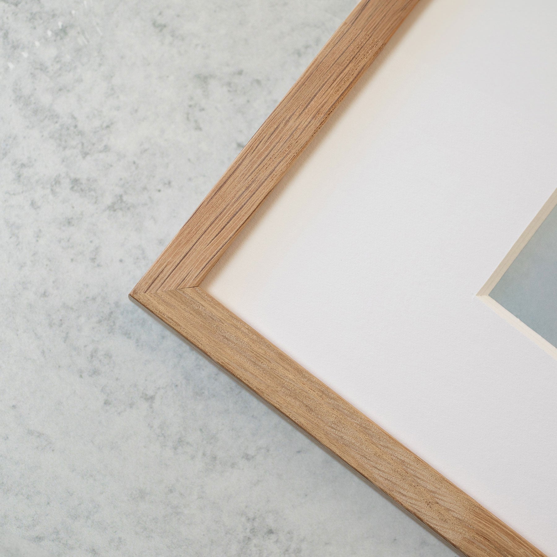Close-up of a wooden picture frame corner against a light gray, textured background. The frame has a smooth, natural wood finish and contains a white mat board. The picture within is printed on archival photographic paper, capturing Seascape Coastal Art, &#39;Surf and Rocks&#39; by Offley Green.