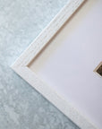 Close-up view of a corner of a white distressed wooden picture frame on a pale grey textured surface, partially displaying a mounted Pink Rose Print on archival photographic paper by Offley Green.