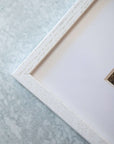 Close-up of a white textured Offley Green picture frame on a gray background, showcasing the corner with a partial view of a photograph on archival photographic paper inside.