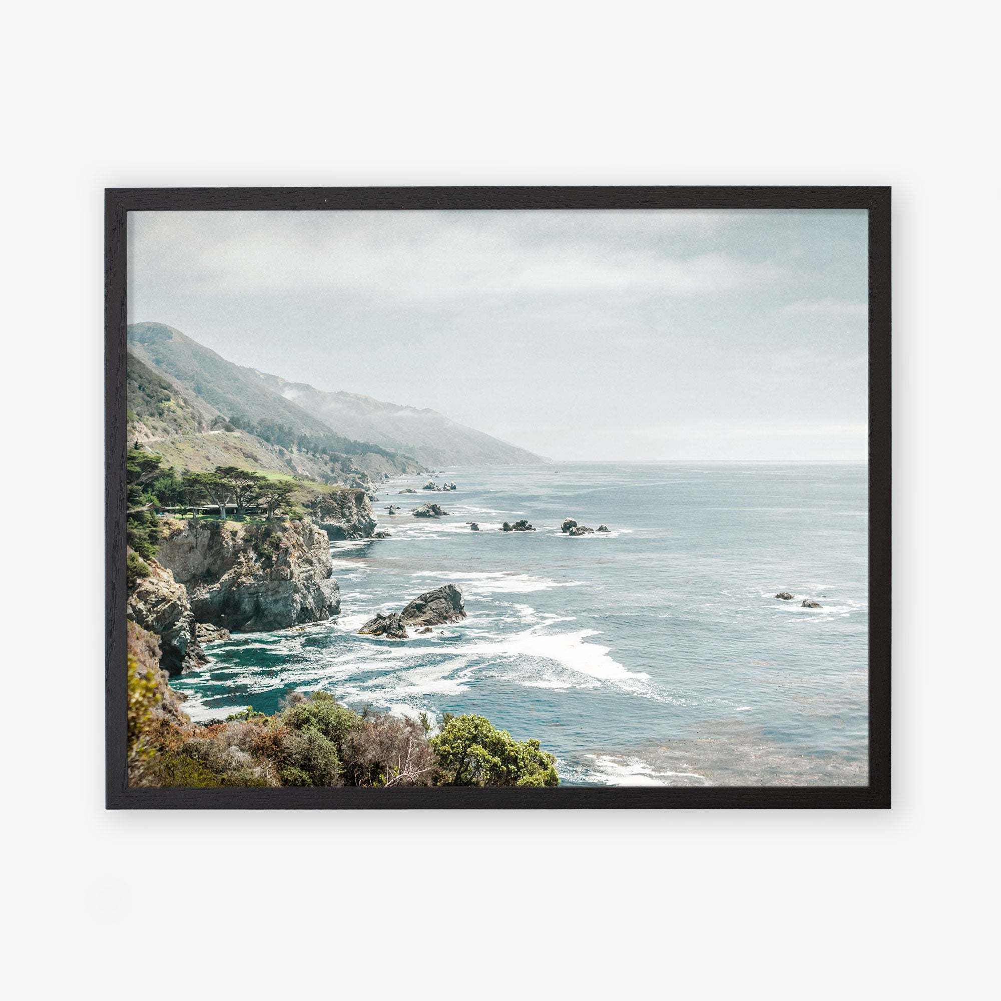 A framed archival photographic print depicting a scenic coastal view of Big Sur with rugged cliffs and a serene ocean under a hazy sky, showcasing the natural beauty of California Highway 1. This is the Offley Green &#39;Rocky Rocks&#39; Big Sur Landscape Print.