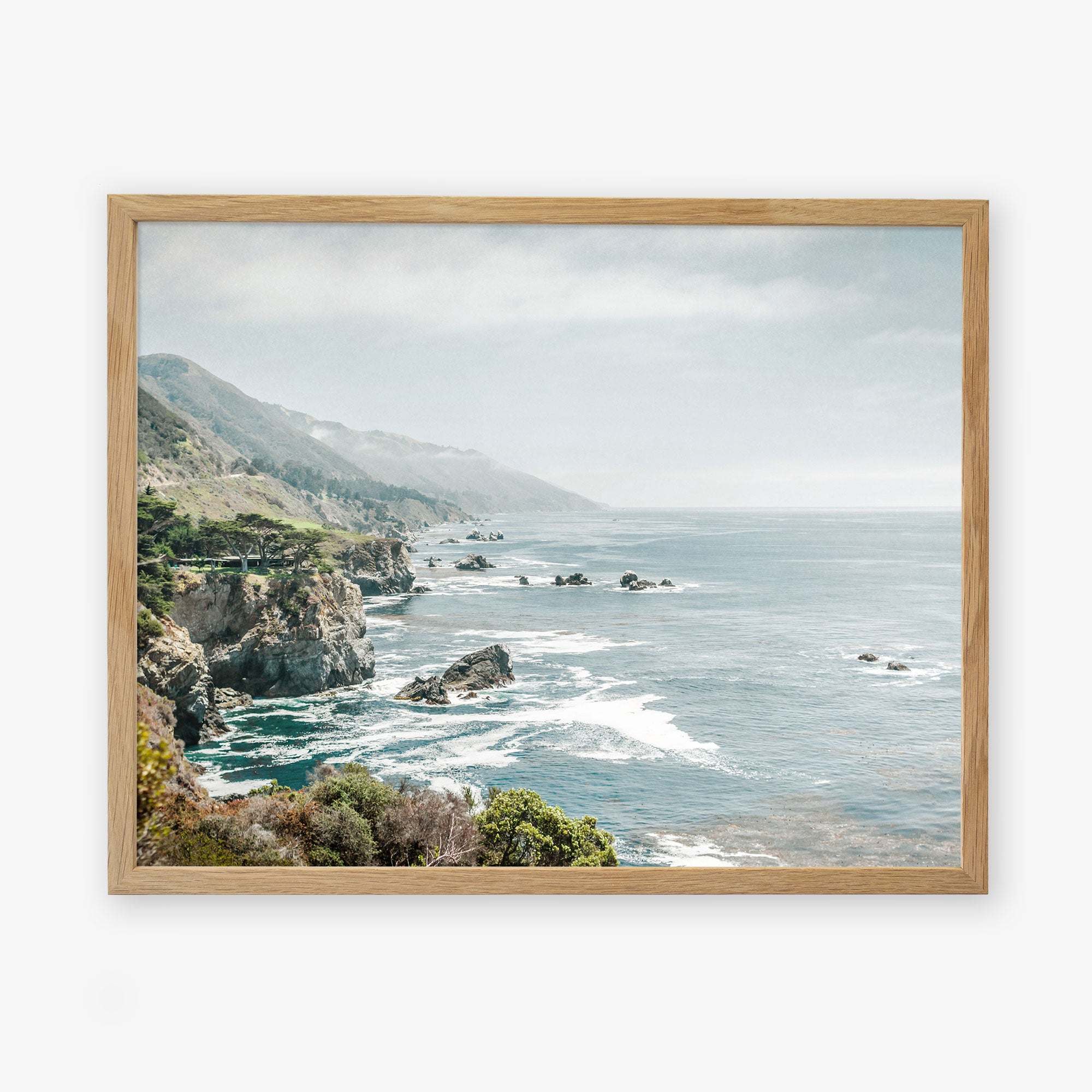 A framed archival photographic print depicting a serene coastal landscape along California Highway 1, with rocky cliffs and a vibrant blue ocean stretching into the distance under a hazy sky. The Big Sur Landscape Print by Offley Green captures the beauty of &#39;Rocky Rocks&#39;.