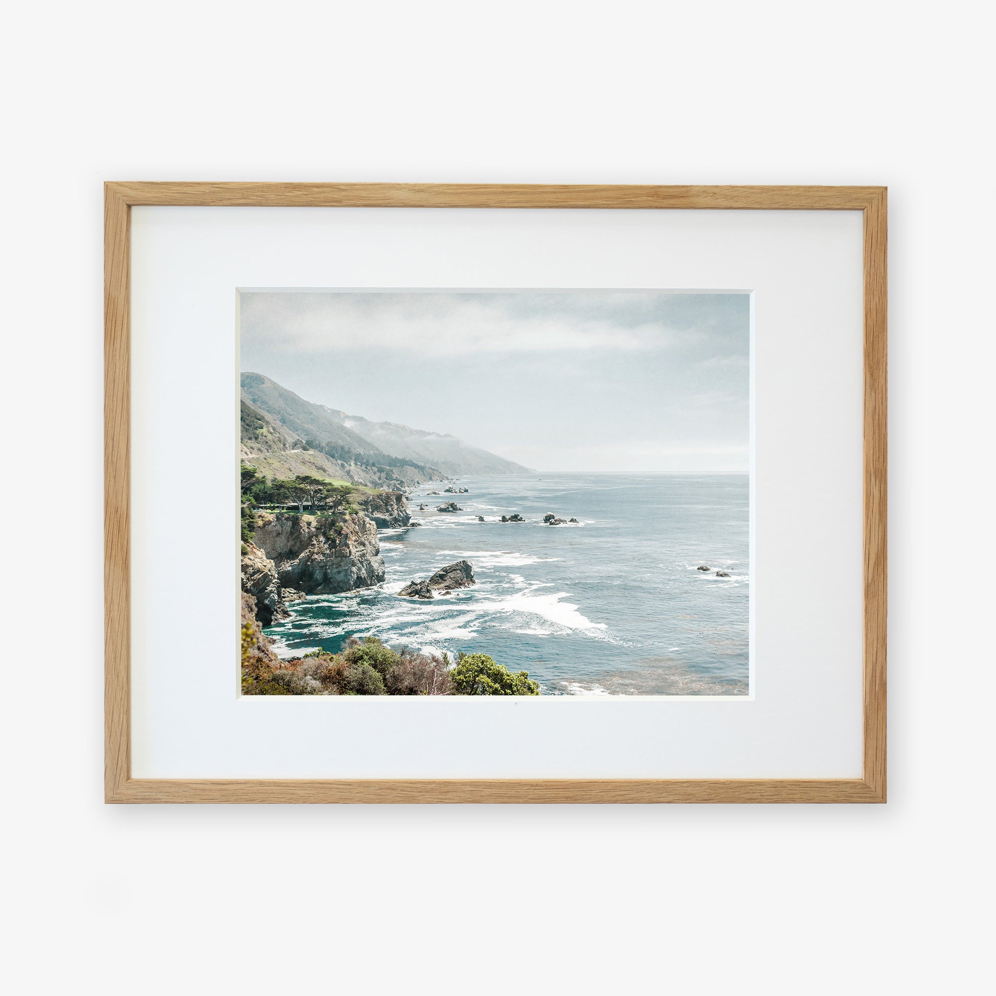 A framed wall art featuring a picturesque coastal scene from Big Sur, with cliffs, the ocean, and distant fog. The light wooden frame complements the serene coastal colors. - Offley Green's Big Sur Landscape Print, 'Rocky Rocks'